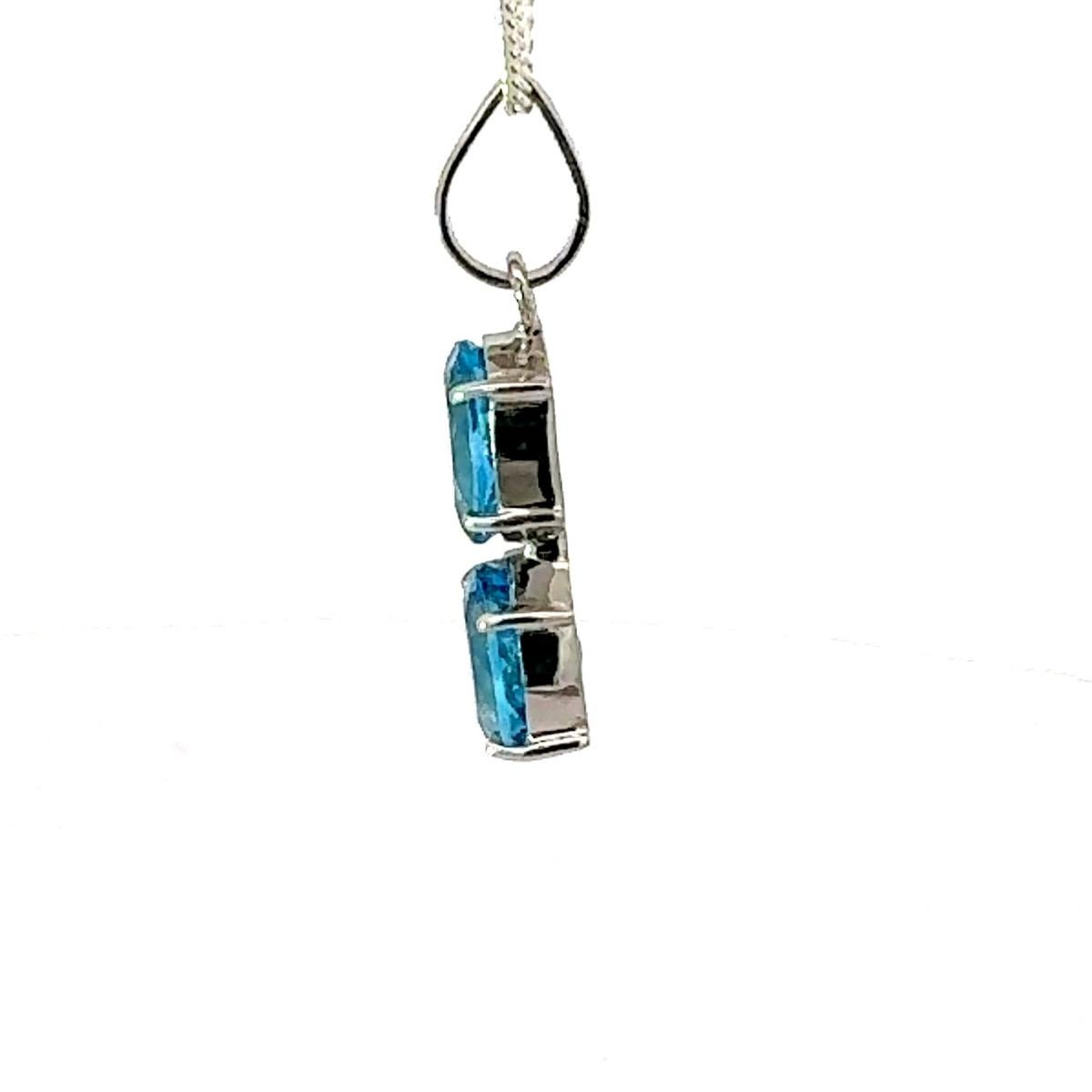 Este colgante de gota vertical con gema gemela de topacio azul es un elegante colgante hecho a mano con dos fascinantes gemas de topacio azul de talla oval de 6,48 cts. Engarzadas verticalmente en un bisel limpio y minimalista, las vibrantes piedras