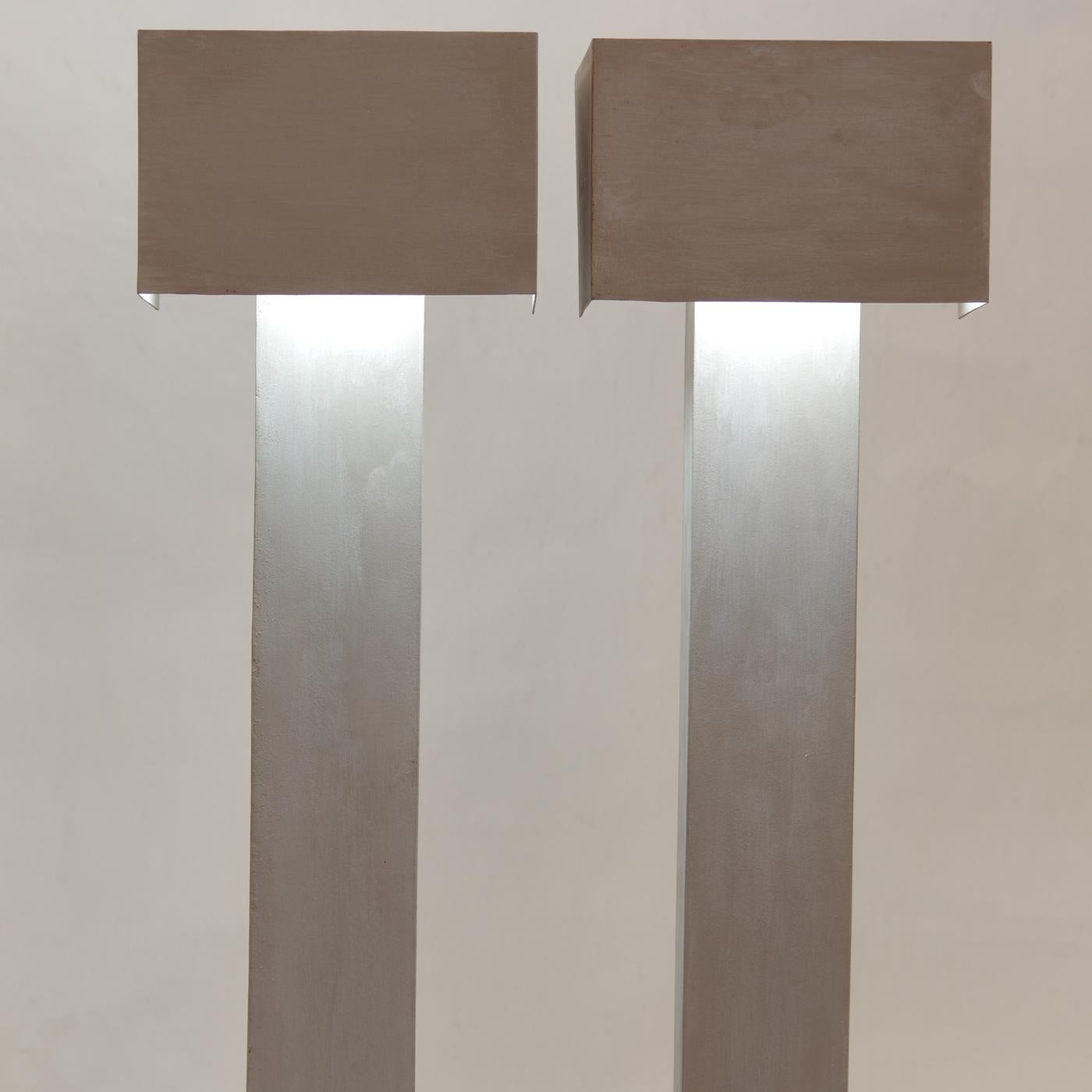 This stunning light sculpture is defined by a T-shaped silhouette, with a polished vertical stem topped by a brown horizontal bar where the light source is nested. The combination of colors and textures is particularly captivating when the lamp is