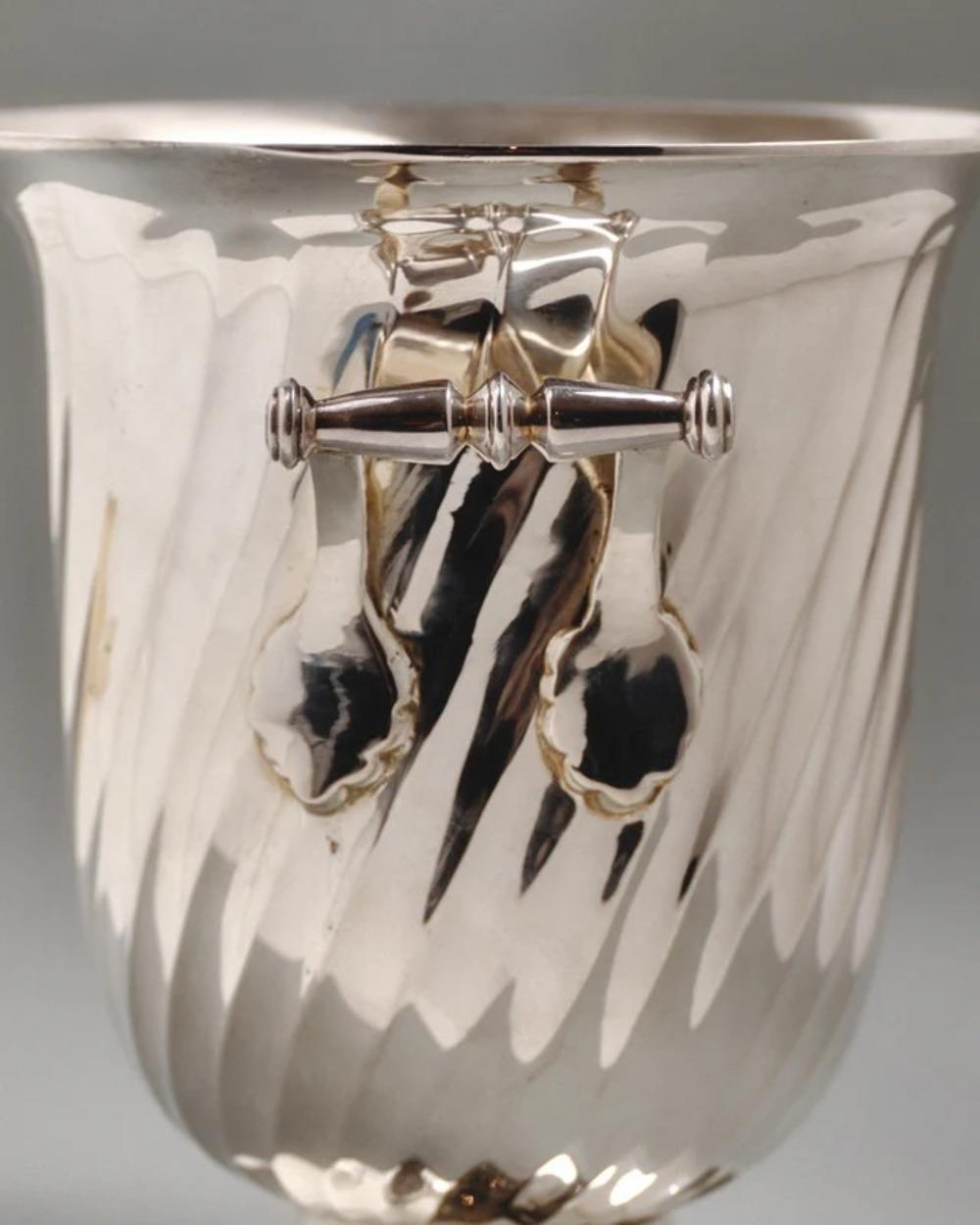 Seau à champagne en métal argenté avec un corps arrondi et cannelé avec des rainures torsadées, reposant sur un pied circulaire. Les poignées latérales sont des barres tournées aux extrémités sphériques. Travaux européens des années 1950. Poinçon