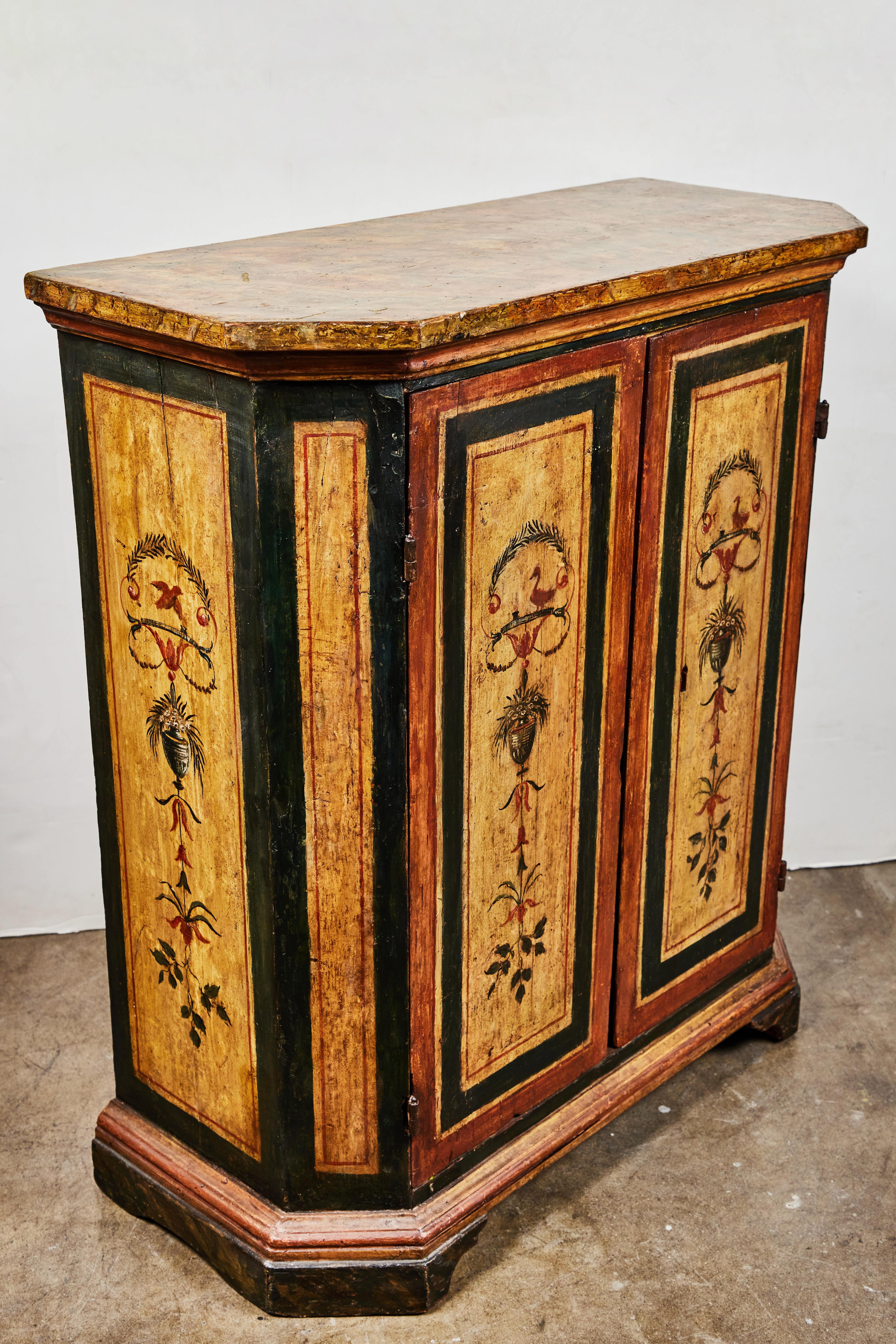 A pair of richly colored, hand carved, polychrome, Italian, clipped-corner, two door cabinets. Each with faux marbled tops and bases. Decorated with vignettes of birds and urns amidst elegant sprays of leaves, flowers, and ribbons. Acquired from a