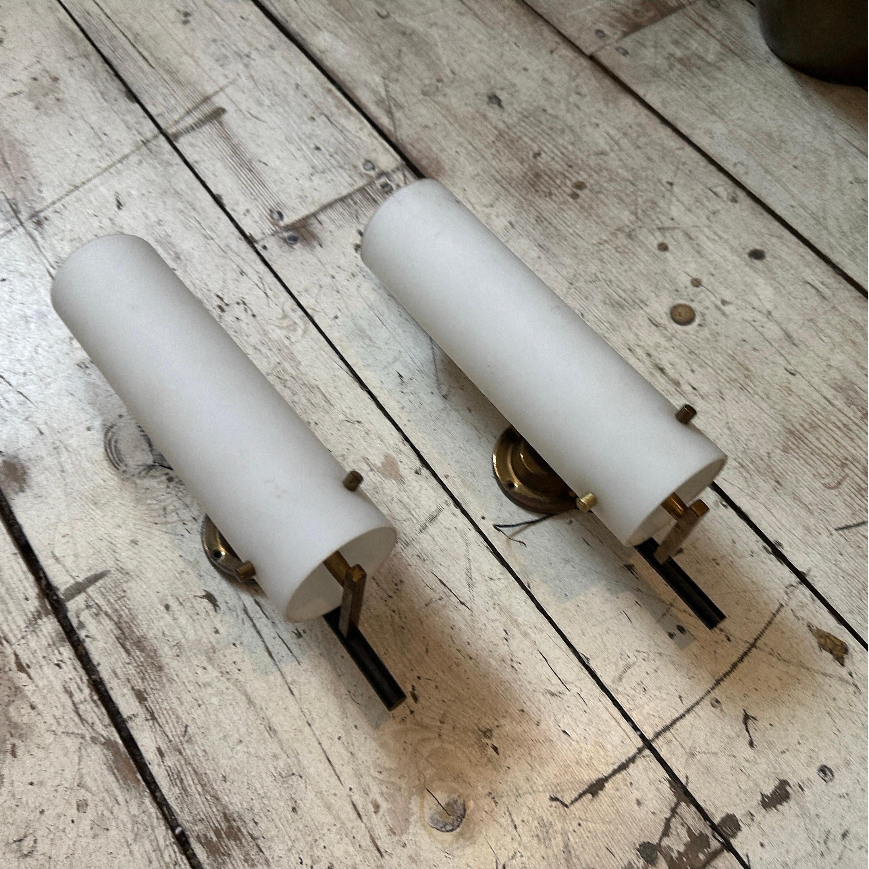 These two wall sconces, crafted in the 1950s and inspired by Stilnovo’s iconic style, are exemplary of Mid-Century Modern Italian lighting design. Each sconce has a cylindrical white opaline glass shade that softly diffuses light for a warm, ambient