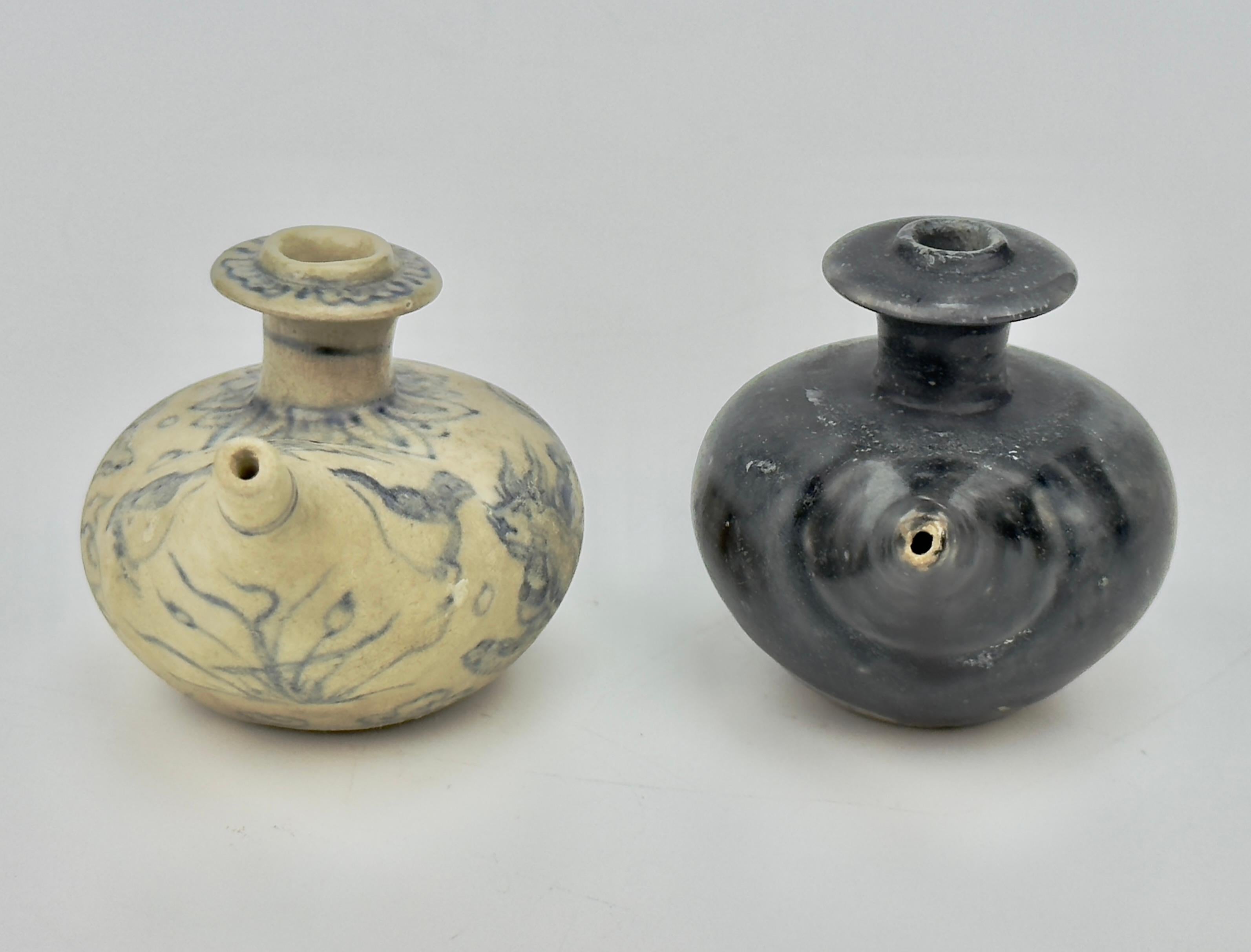 These 15th-century Le Dynasty ceramic vessels from northern Vietnam feature distinct designs. One is beige with faint blue floral patterns, a rounded body, and an elongated spout. The other is navy, undecorated, with a similar shape and a short,
