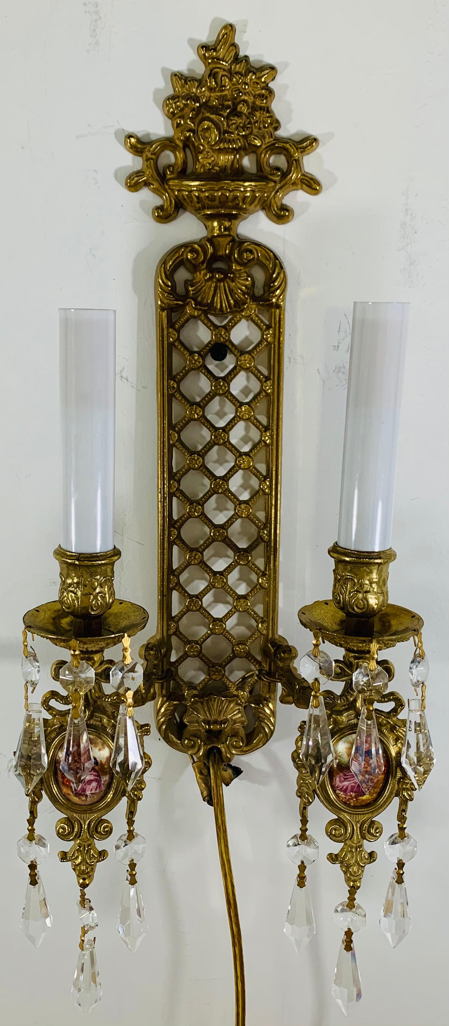 A pair of beautiful brass wall sconces in the style of Louis XVI. The pair features faux royal Vienna print oval inserts with three beautiful crystal prisms hanging from each insert. Each sconce has two arms with candelabras. 

Measures: 6.75 W 17