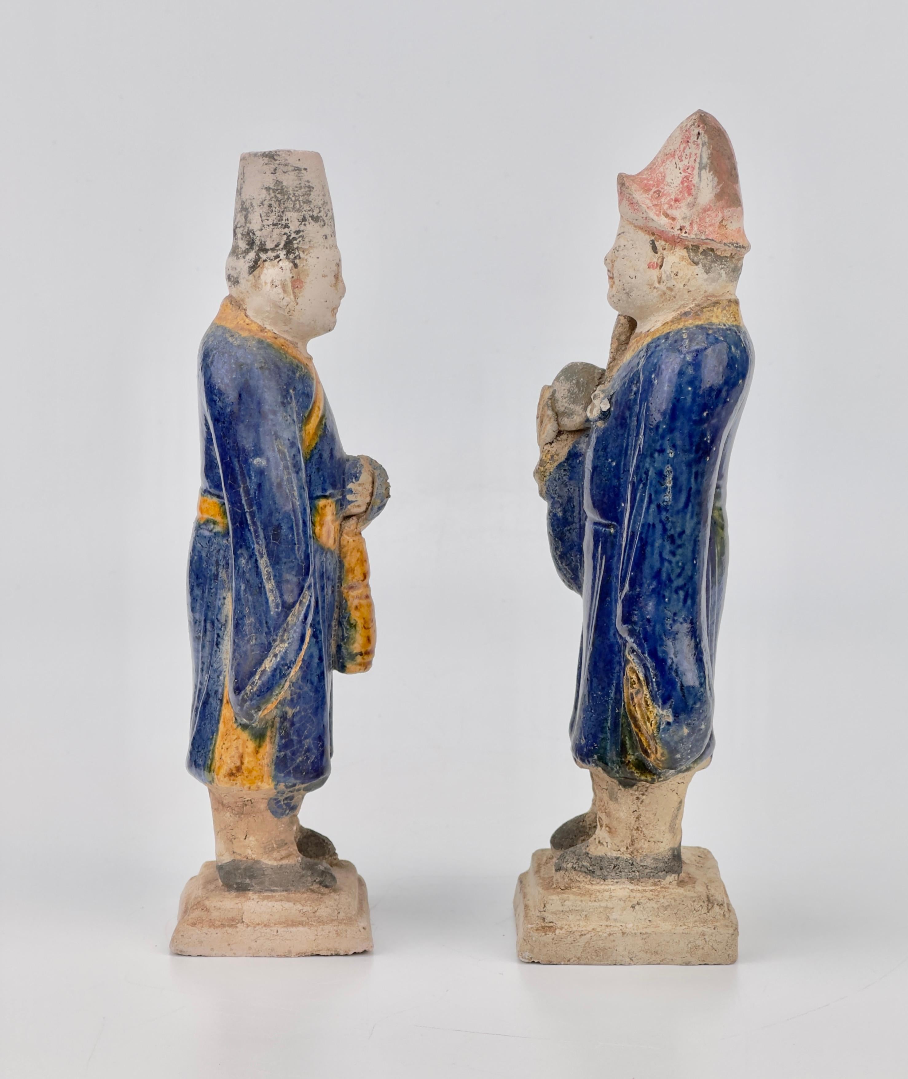 Statues of Chinese dignitaries crafted from terracotta, featuring glazes in blue and ocher, are set on rectangular bases. The wide-sleeved robes and craftsmanship, along with the cylindrical hats, indicate their origin, specifically from the Ming