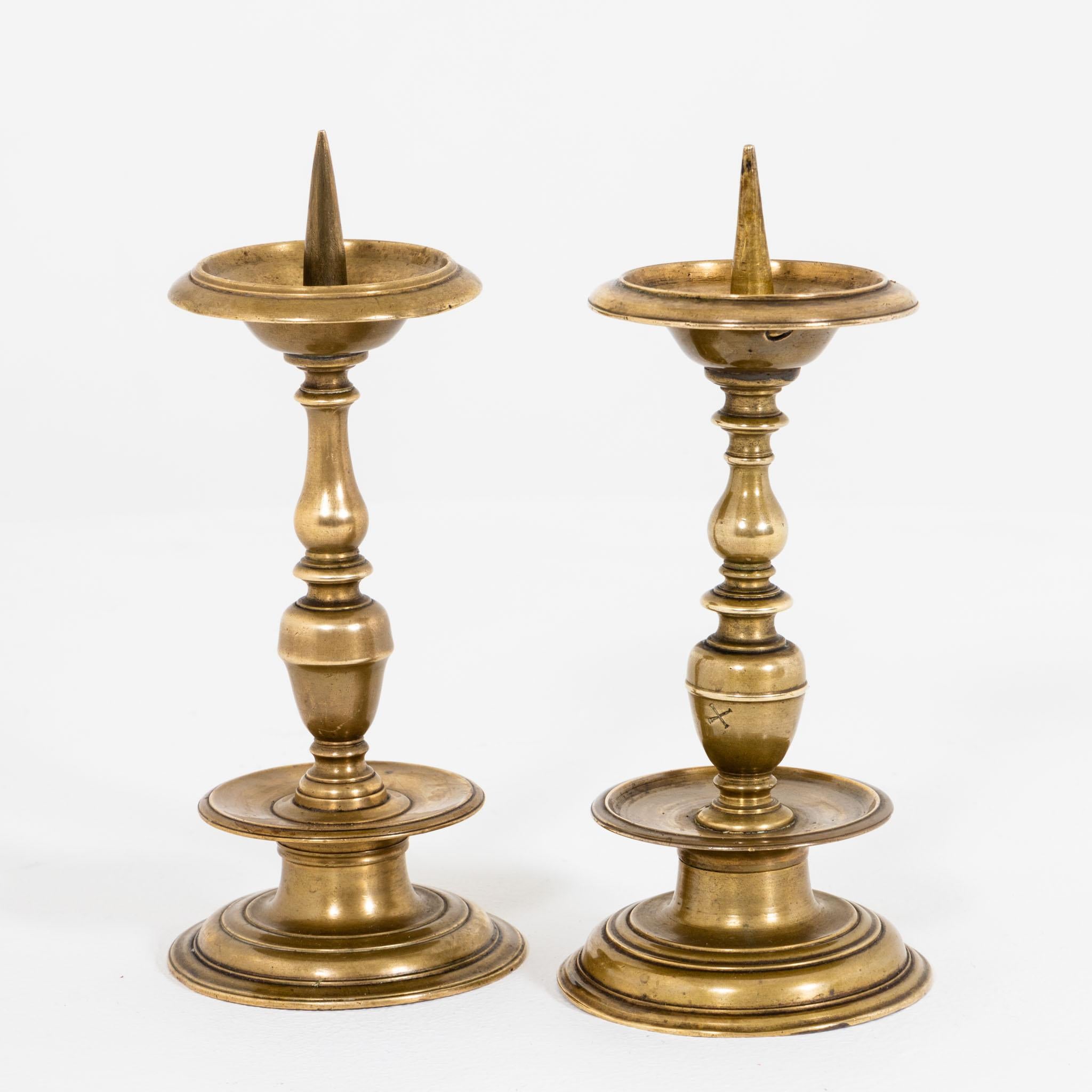 Two brass candlesticks with balustraded shafts and drip trays with spikes. The candlesticks are not identical in their design, but complement each other decoratively to form a harmonious ensemble. One with a small hole on the drip tray and X marking.