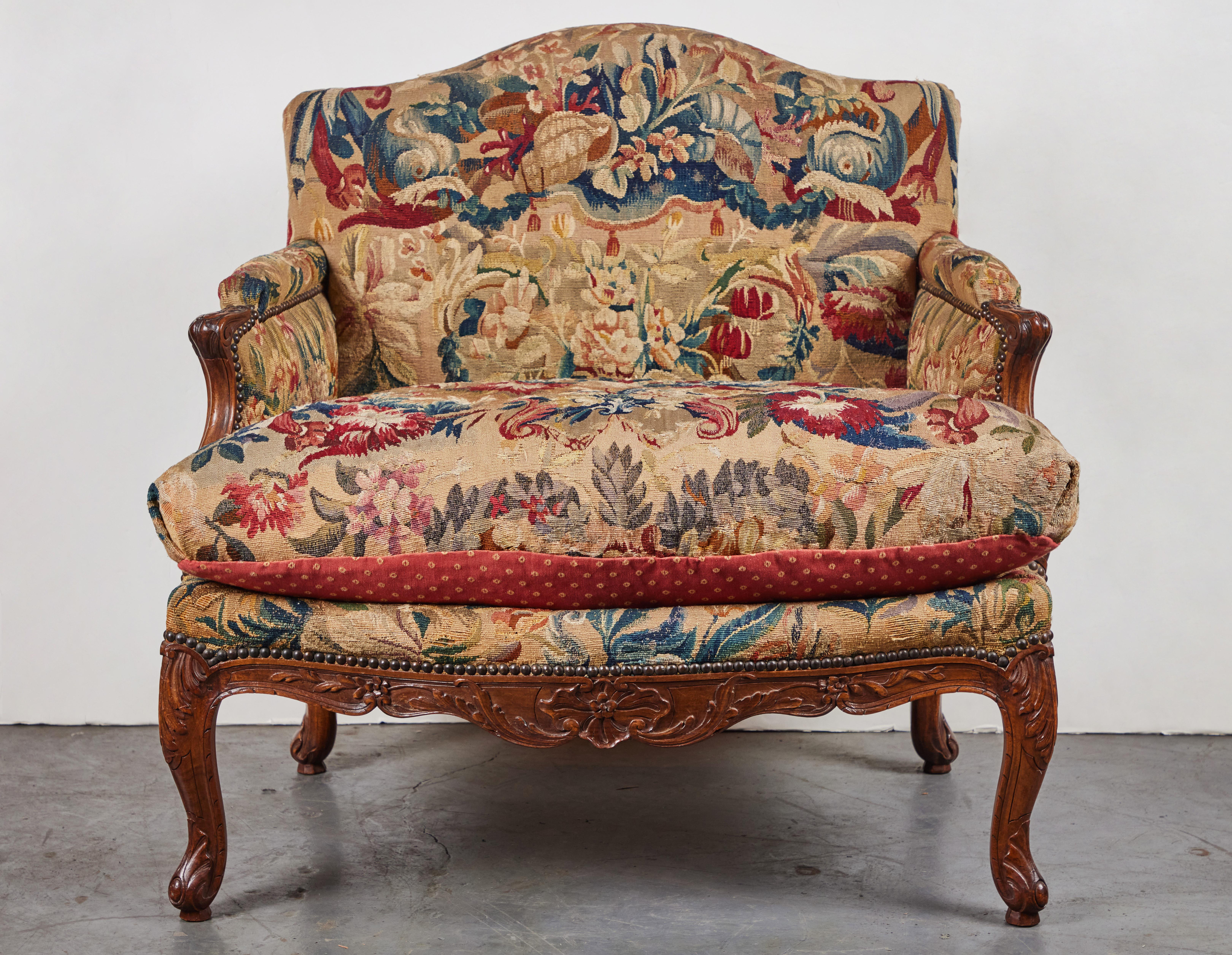 An exceptional pair of period, hand-carved walnut, Provencal marquis chairs in dramatic hand-stitched, tapestry style fabric featuring flowers and leaves affixed by bronze nail heads throughout. The apron and arms embellished with foliate relief