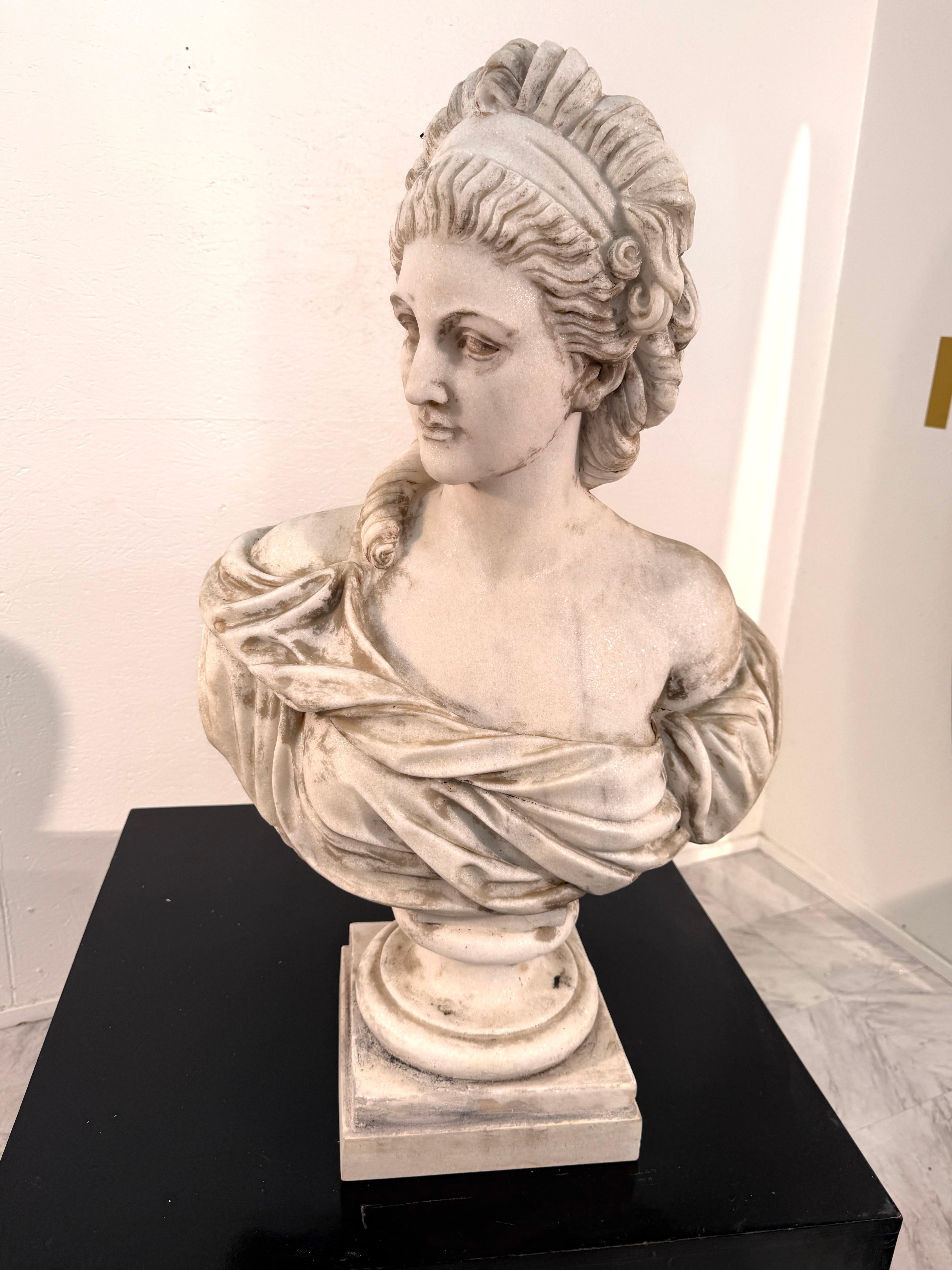 A graceful white marble bust depicting a refined classical lady, draped in softly carved garments and adorned with an elegant headpiece. The sculpture captures serene beauty and timeless sophistication, with intricate detailing in the cascading