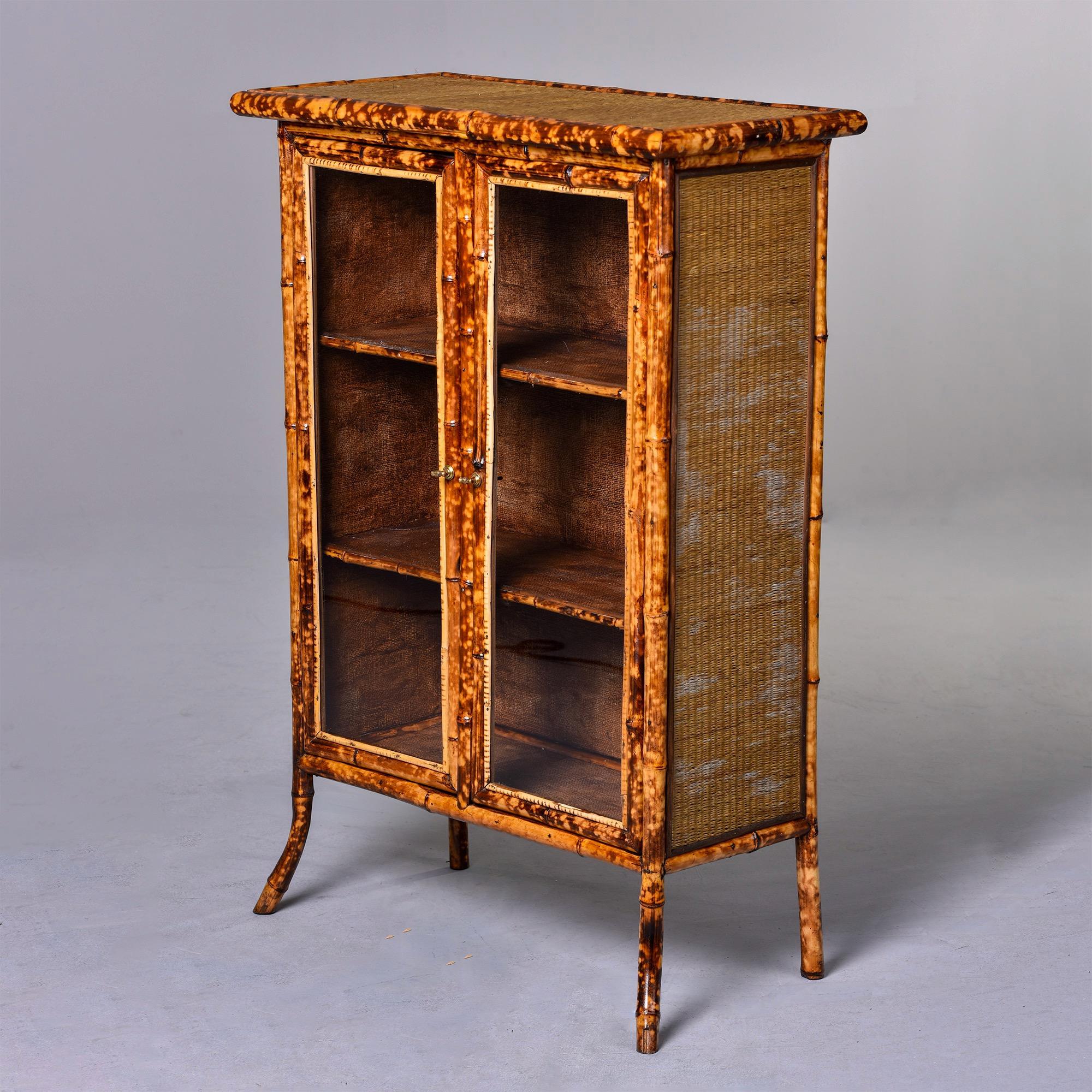Circa 1930s two door spotted bamboo cabinet features glass fronts with woven grass cloth sides and top and two interior shelves. Unknown maker. Found in England.