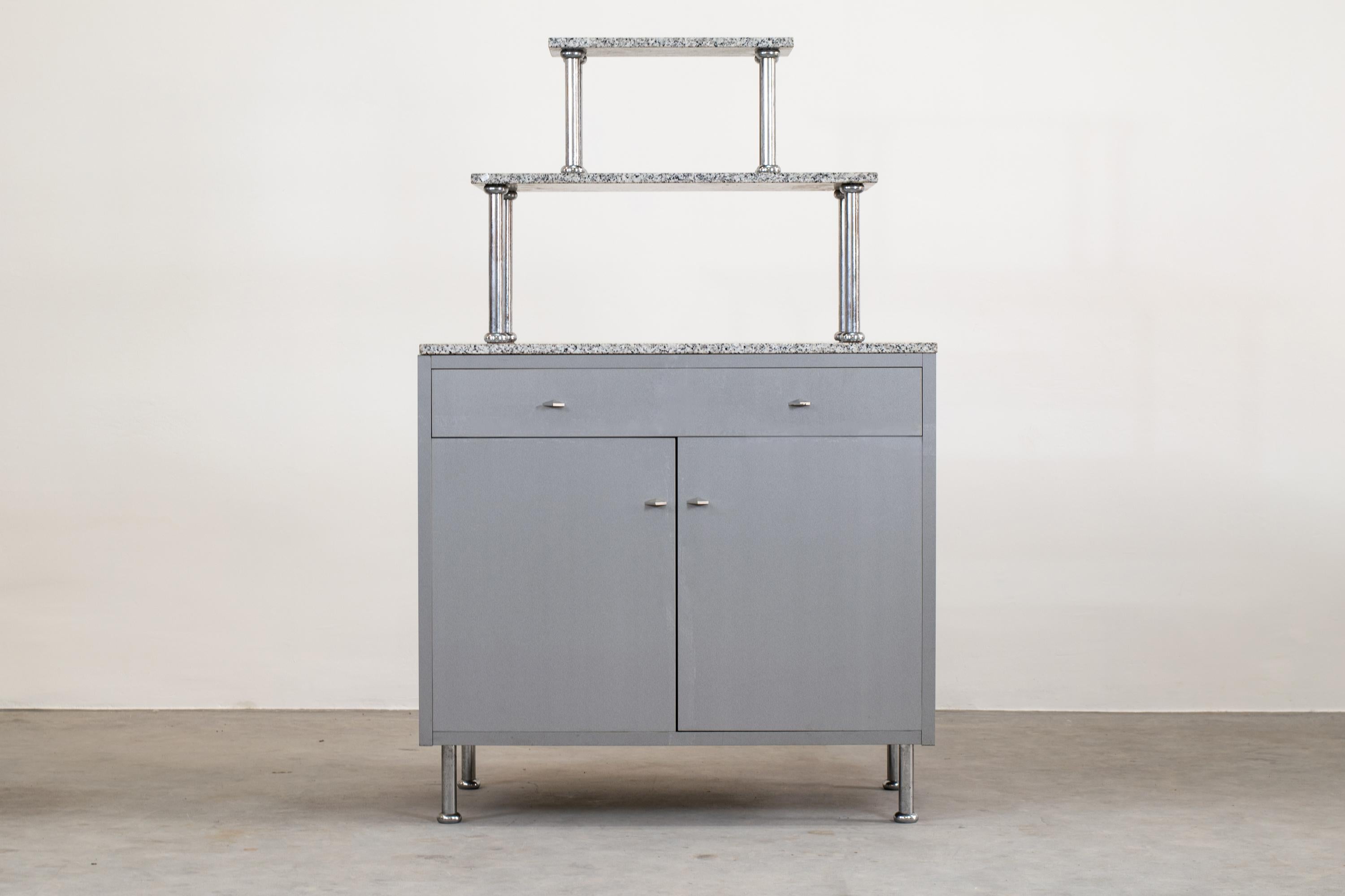 Cabinet with two doors, inner shelves, and drawers from the Afrorismi series, it's made in gray metalized and grooved laminate, with legs and upper support in chromed metal, and overlapping shelves in Baveno granite.

Produced by Driade in 1984,