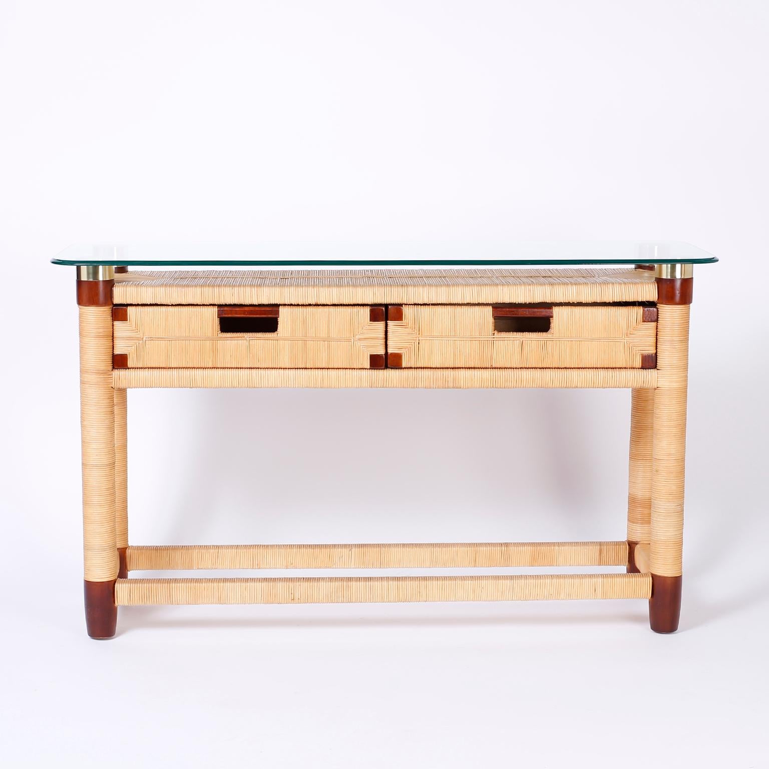 Two-drawer beveled glass top console table by Donghia, with a sleek modern form, a wood frame wrapped with reed, brass capped posts, and mahogany accents including drawer handles and turned feet.
