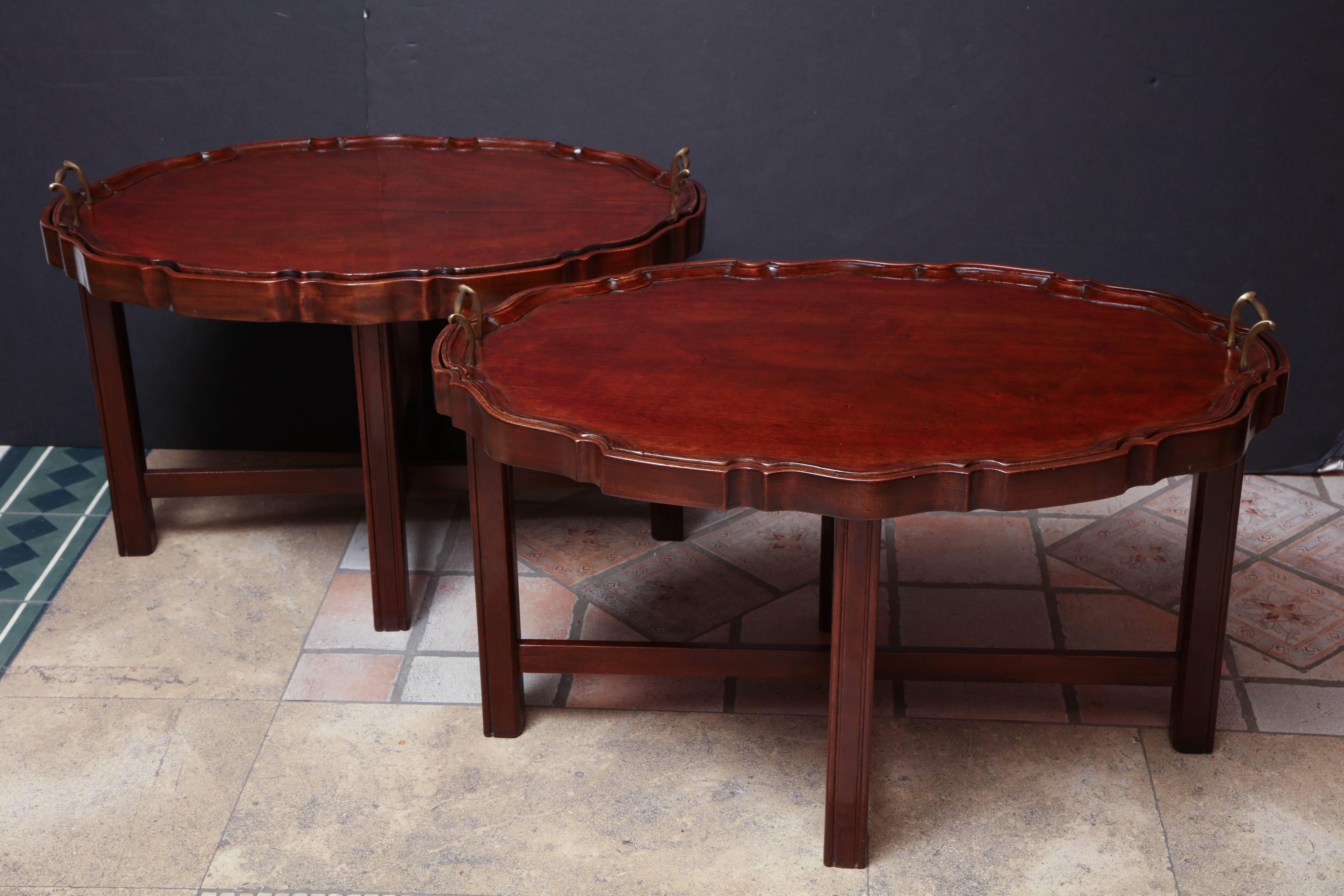 Two English mahogany oval pie crust trays on later molded mahogany stretcher base stands. Each tray with a carved molded edge and brass handles.