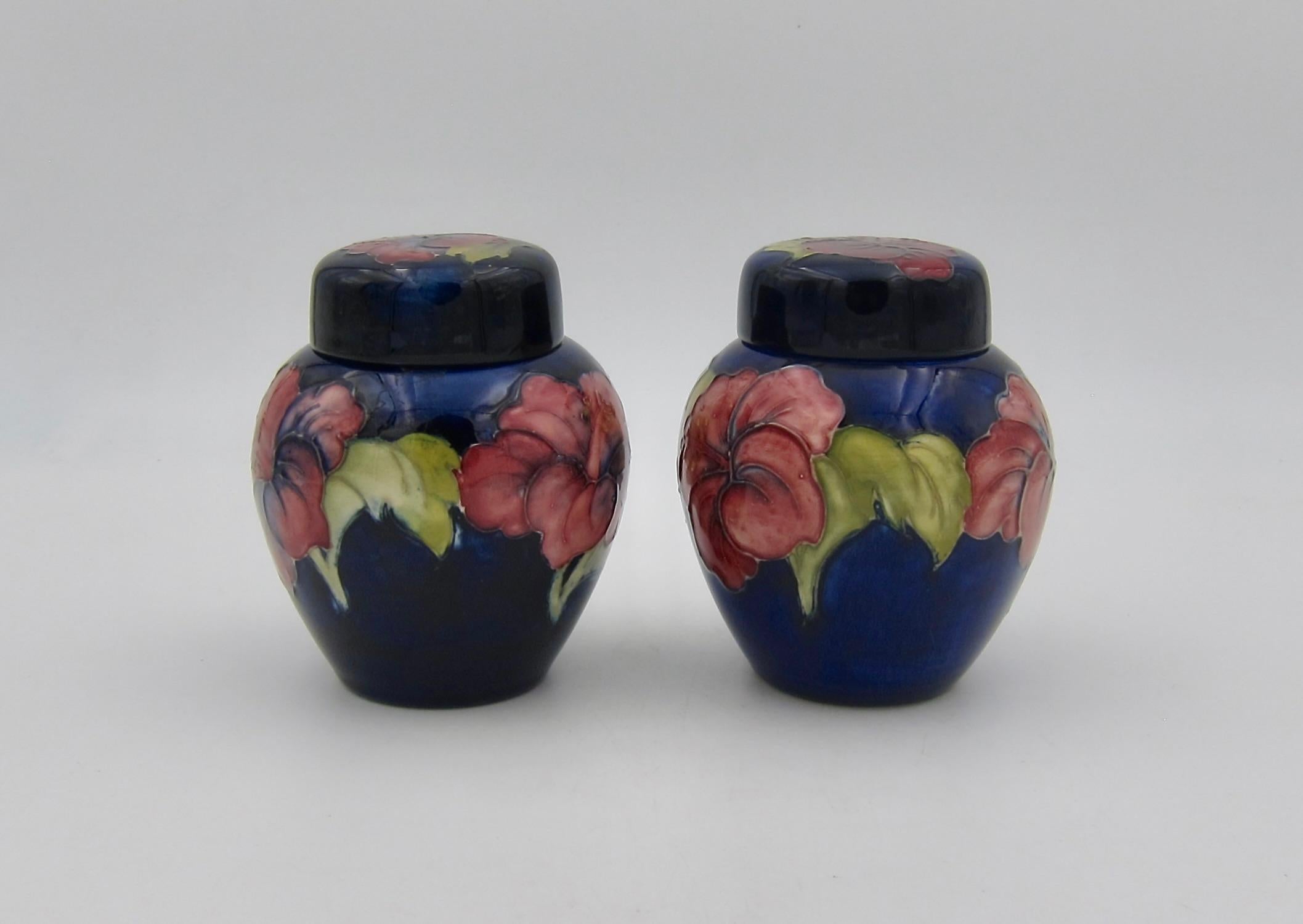 Two vintage Moorcroft art pottery ginger jars in the hibiscus pattern, manufactured at the Moorcroft Pottery at Stoke-on-Trent in Staffordshire, England. Each two piece jar features a ring of colorful tube-lined, hand-painted hibiscus blossoms in