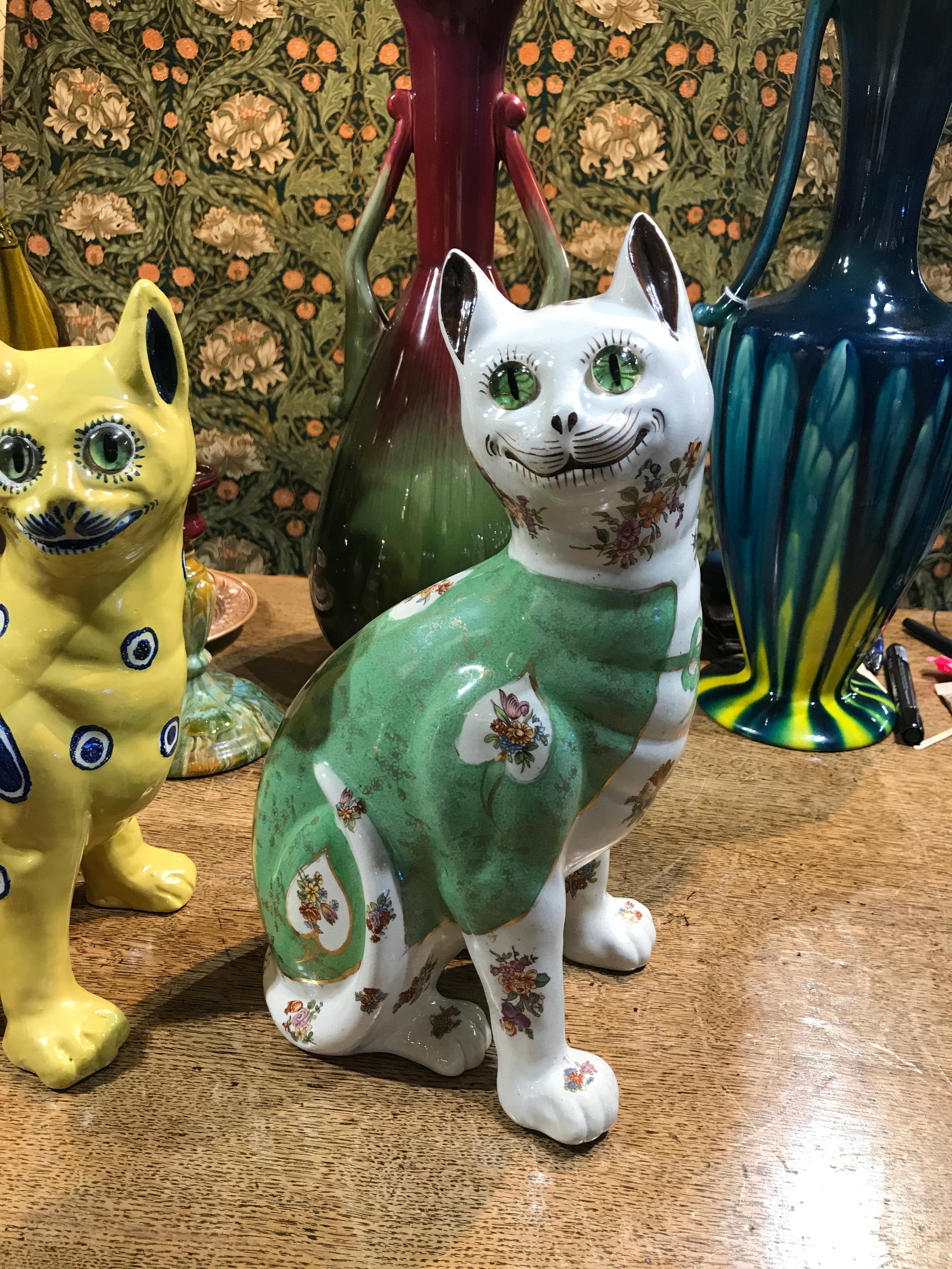 Two French Masonic pottery figures of cats with glass eyes, height 12'.
One with yellow body decorated with blue and white hearts and stylized fish.
One with a pale green body with white panels decorated with sprays of flowers.

 