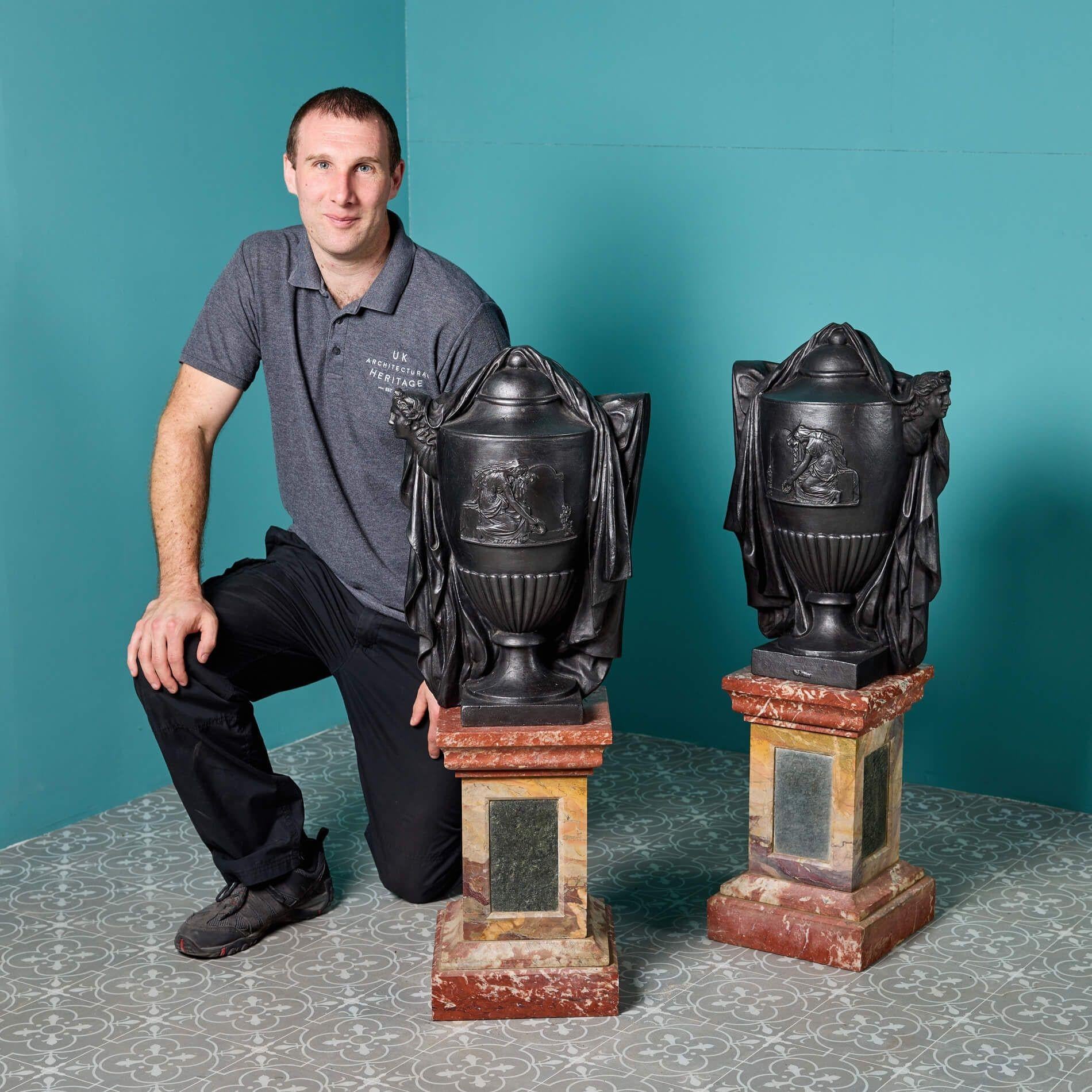 pair of late 19th century French neoclassical style cast iron urns on raised rouge marble pedestals. Each urn is of lidded form and beautifully cast as if draped in fabric with a matching neoclassical depiction of a figure to the front and handle.