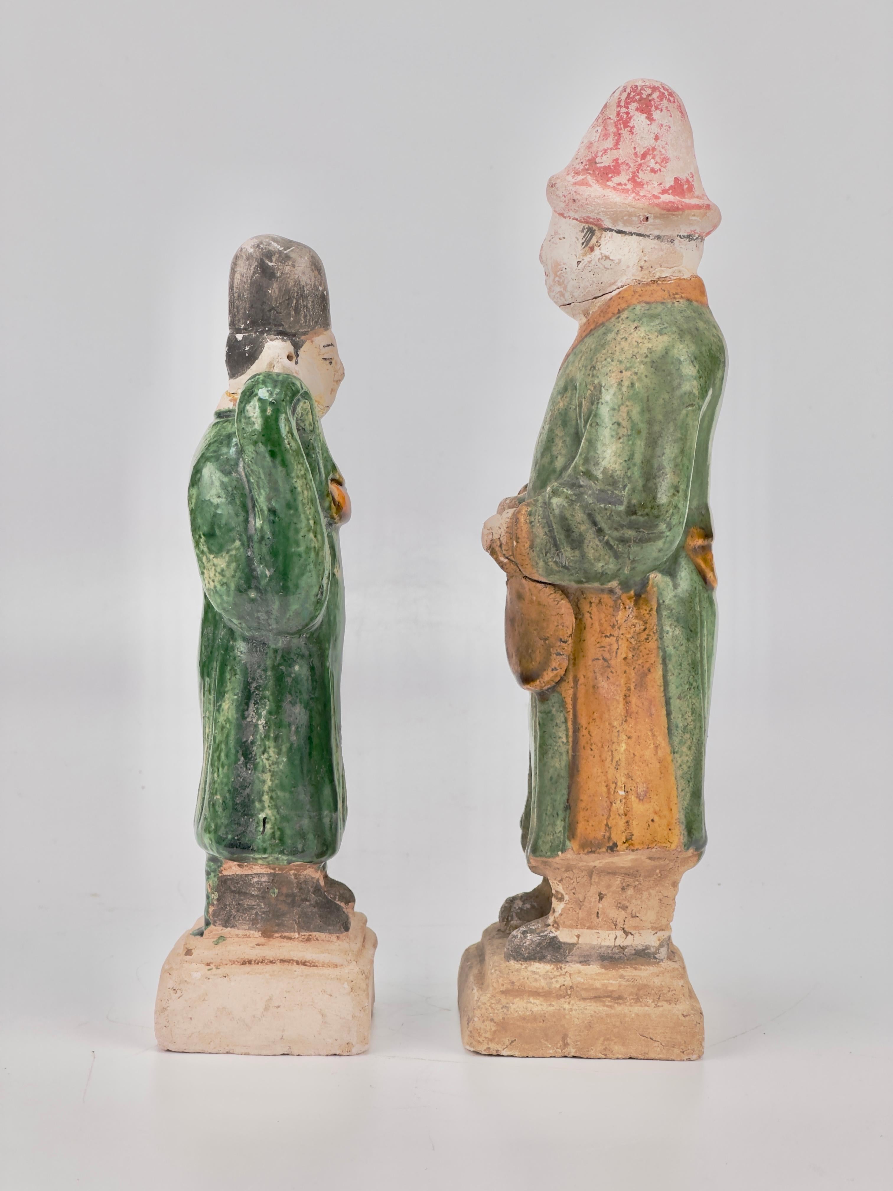 Statues of Chinese dignitaries crafted from terracotta, featuring glazes in green and ocher, are set on rectangular bases. The wide-sleeved robes and craftsmanship, along with the cylindrical flat hats, indicate their Chinese origin, specifically