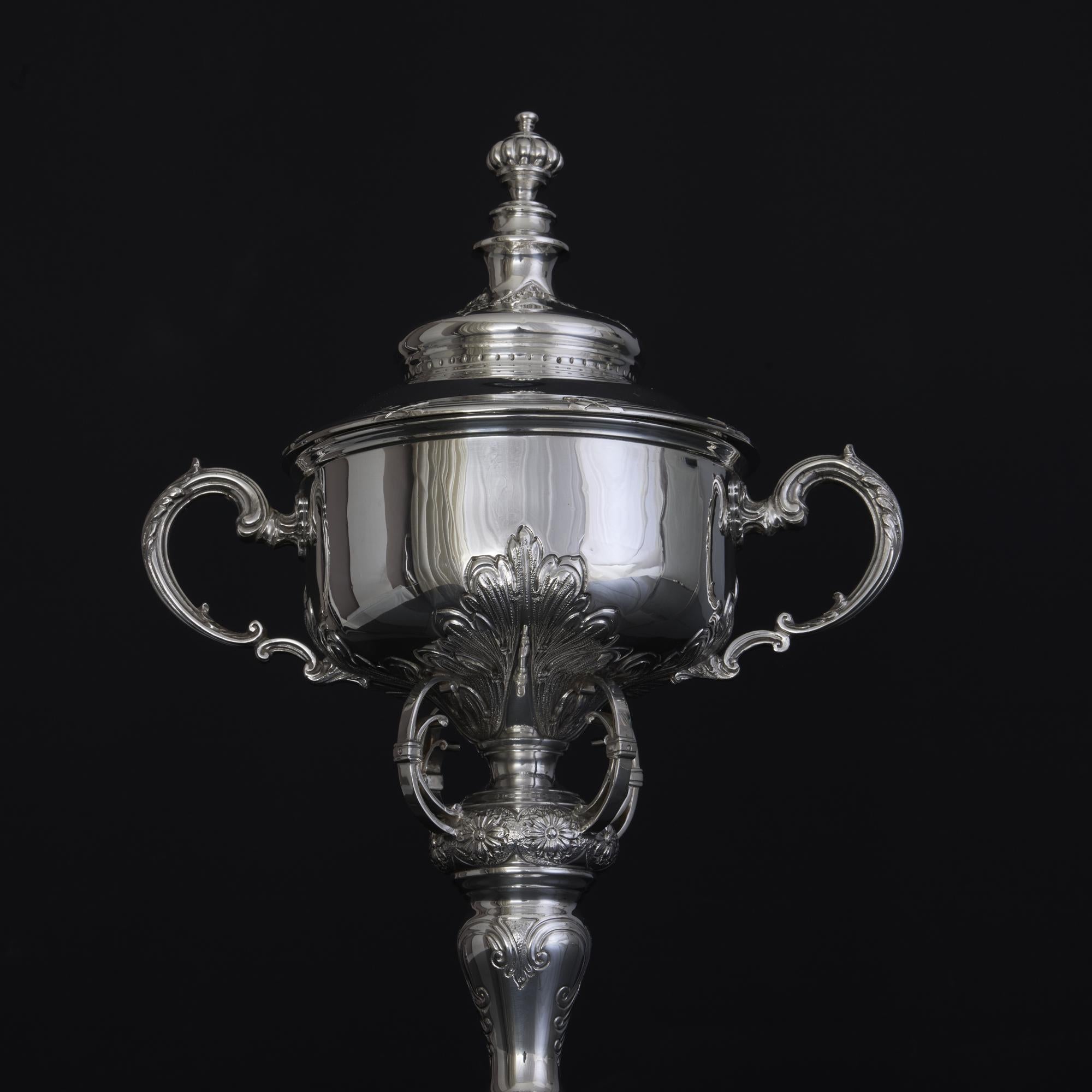 Highly decorated silver trophy cup and cover with hand chased leaf and flowerhead decorations, open-work supports and reed-&-ribbon pattern mounts around the cover which, in turn, is surmounted by a fluted finial.
 The elegant handles are similarly