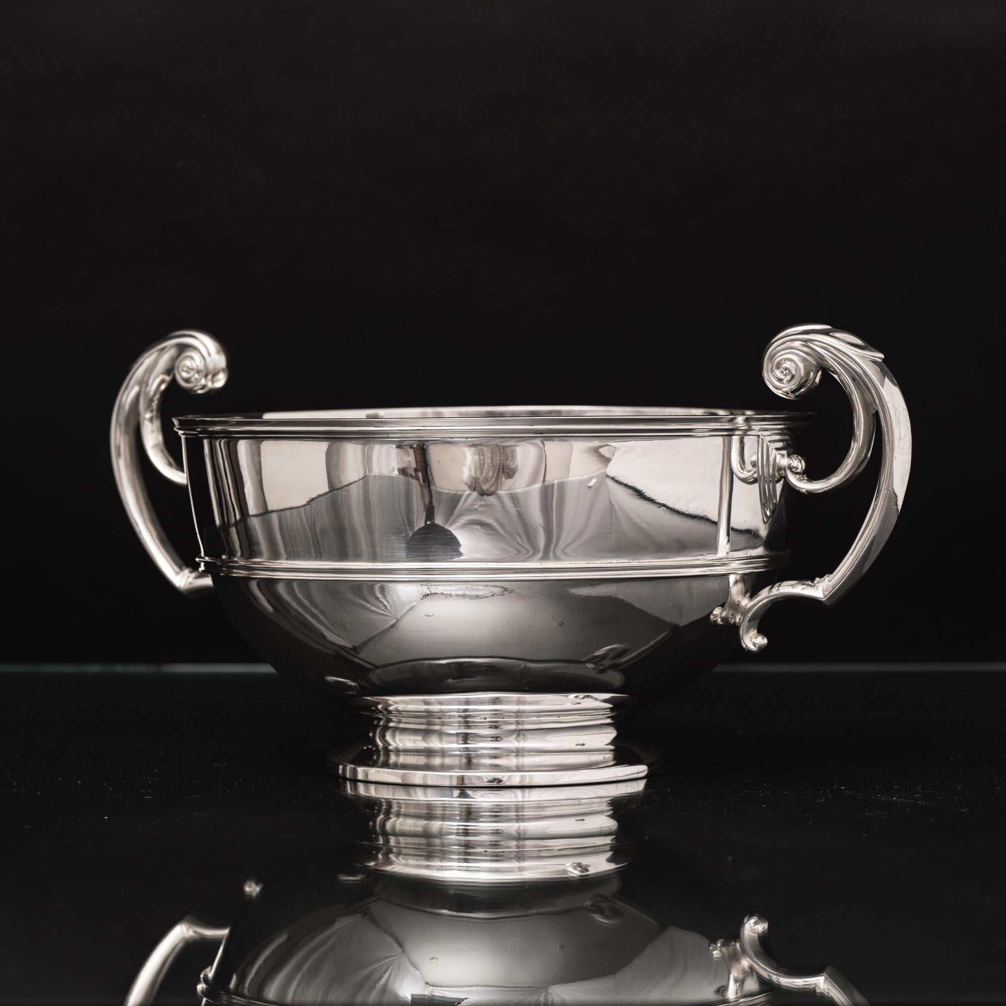 Traditional antique circular silver rose bowl mounted on a spreading circular base and featuring two c-shaped, flying-scroll, raised handles. A reeded girdle encircles the body of the bowl and an applied thread pattern around the circular spreading