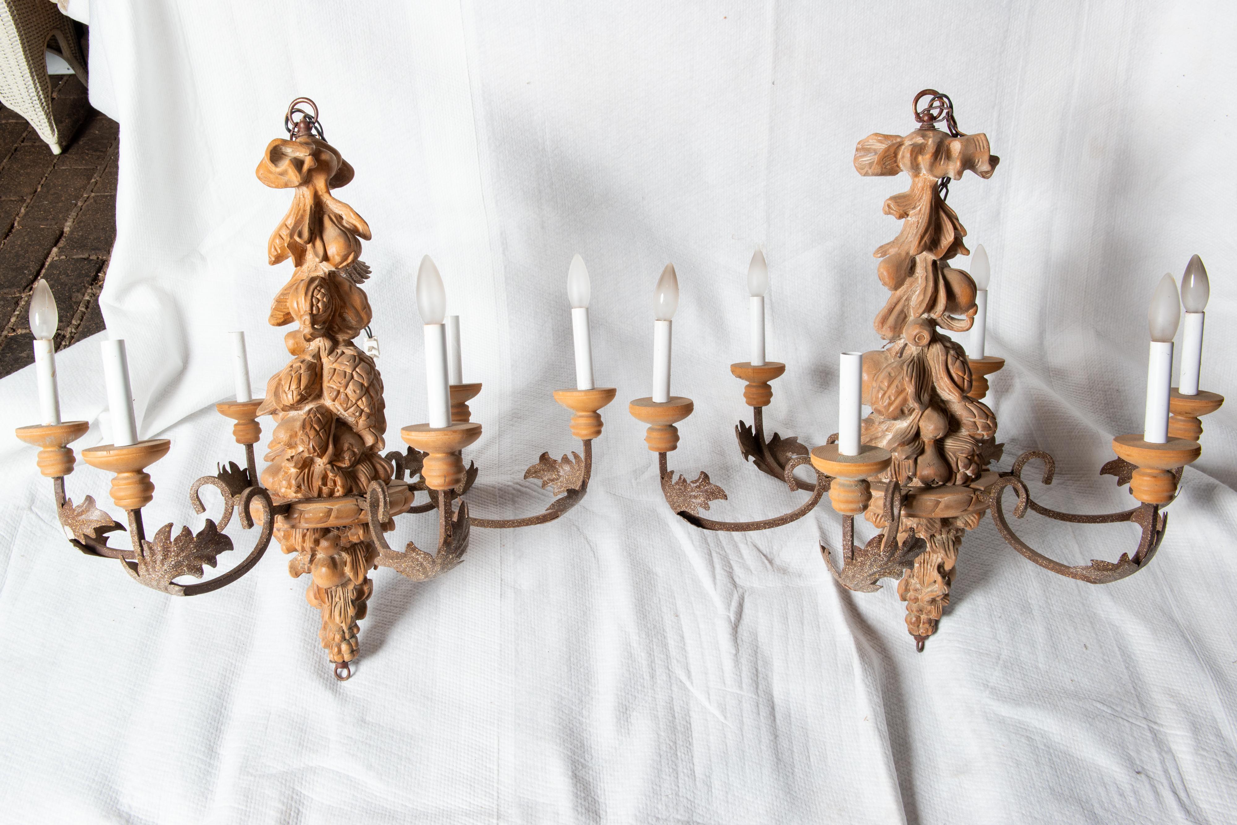 Due disponibili. Lampadario italiano in legno intagliato e ferro battuto. Le sculture in legno includono frutta e fogliame. I delicati bracci in ferro battuto sono ornati ciascuno da una foglia in ferro battuto. Ogni lampadario è dotato di sei