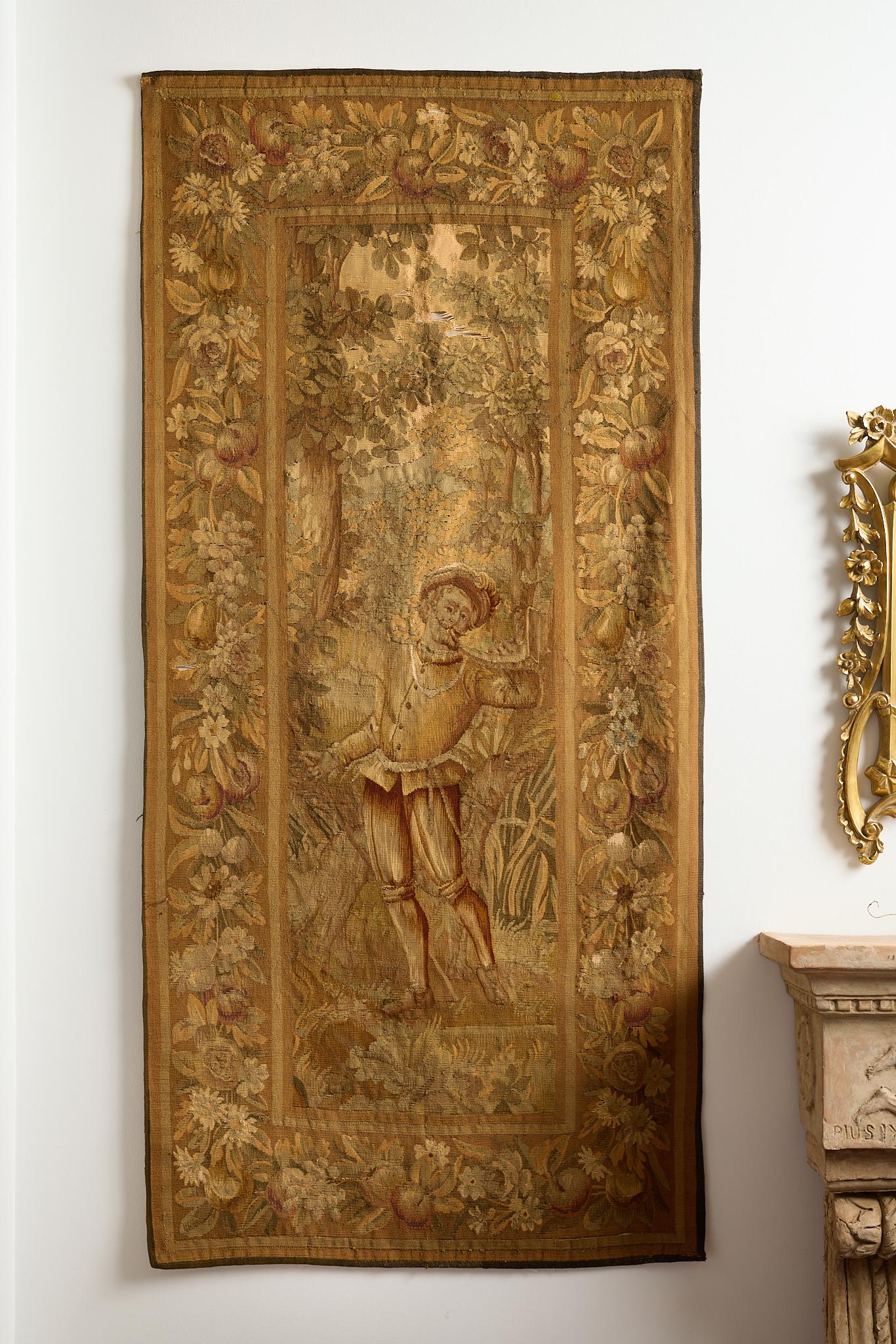 Two large, 18th century, French figural tapestries, each with a central male figure standing in nature, one blowing a hunting horn and one with his arm raised in a searching gesture. Both with a frame of fruits and flowers. 
These tapestries have