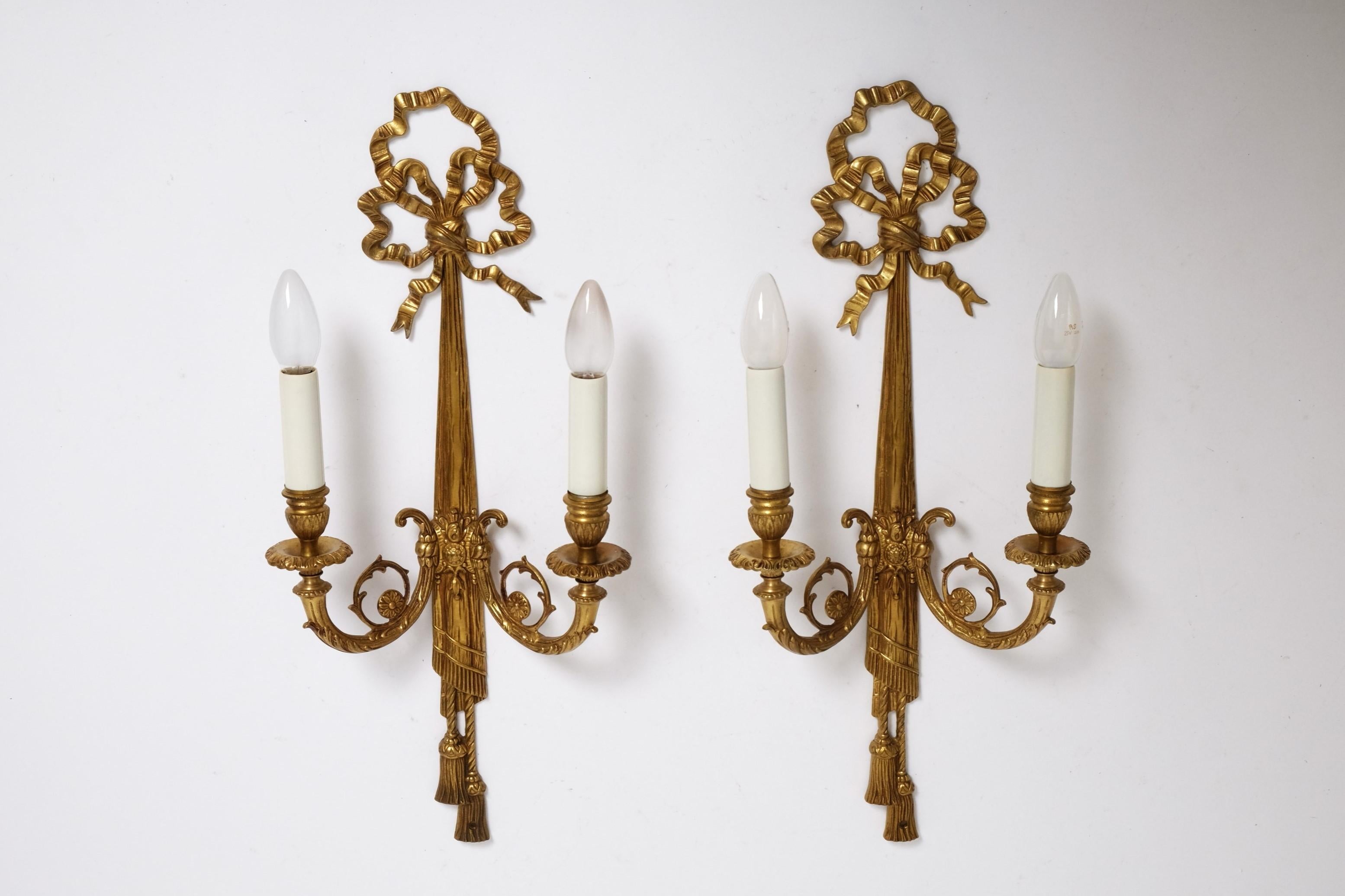 A large pair of French Louis XVI Style Sconces from the 1950s. Twin-armed ‘grand appliques’ in cast bronze with elaborately decorated arms and chased detailing. Decorated with bows, drapes and trim. Each lamp has two light arms decorated with