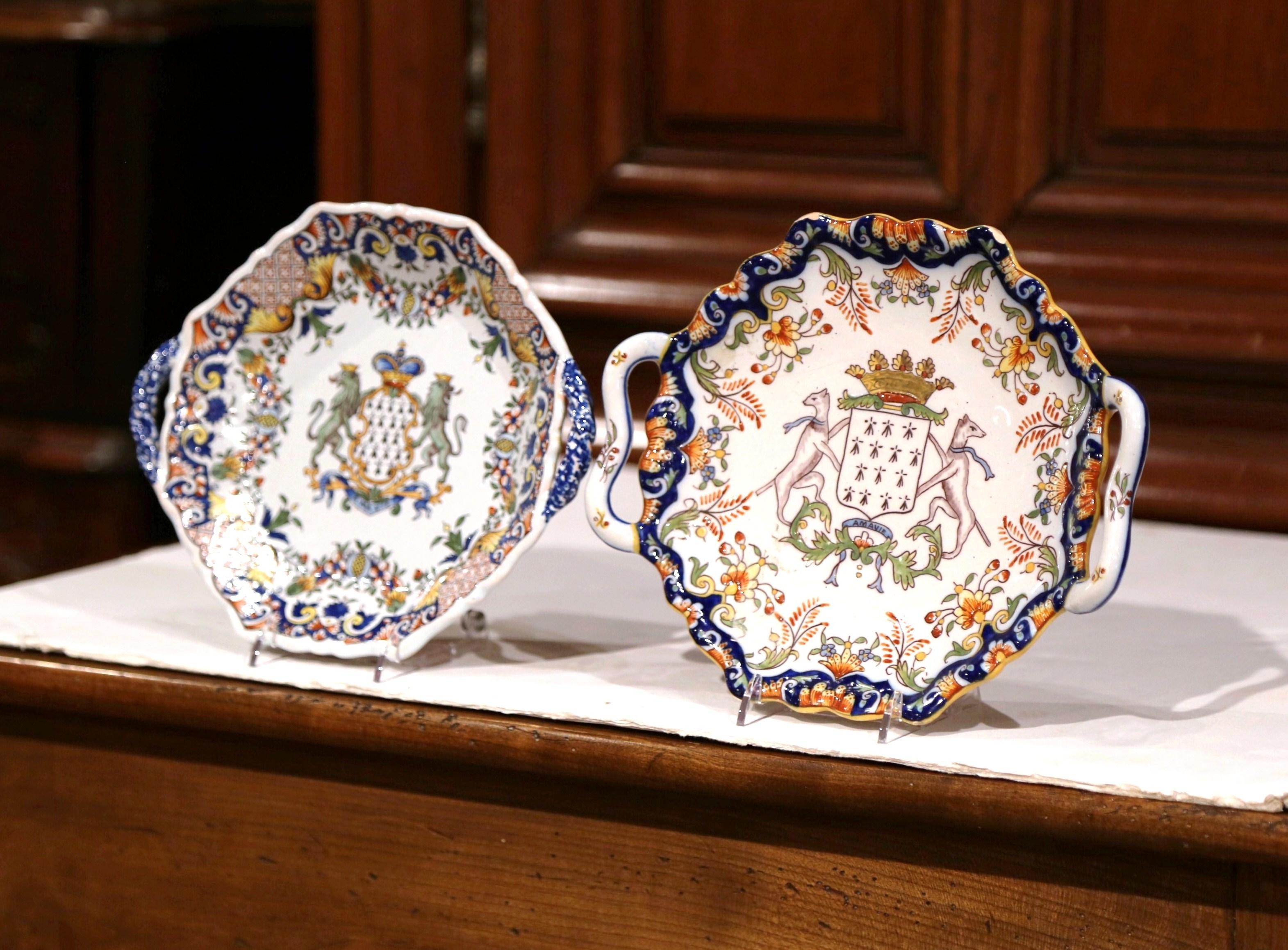 Decorate a shelf or a wall with these colorful antique decorative plates from Brittany, France. Crafted circa 1930, each faience plate has handles and features a hand painted centre crest. The plates are further embellished with floral decor around