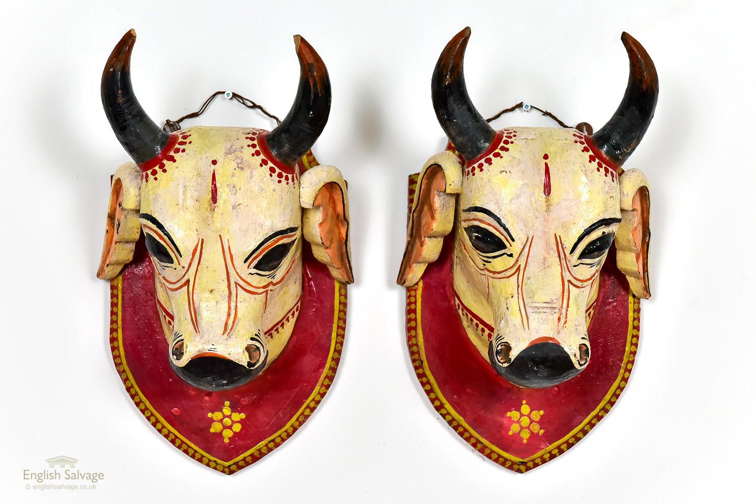 Two carved cow heads, brightly painted and mounted on carved shields to resemble trophies. They both have a hanging wire connecting to two lugs to support them. One has some damage to its horn and both have some chips and cracks as expected from