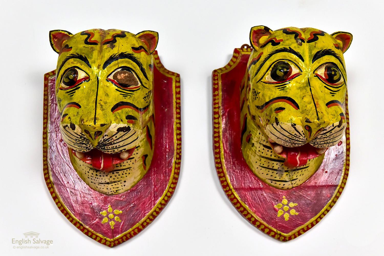 Two uniquely carved tiger heads, brightly painted and mounted on carved shields to resemble trophies. They both have a hanging wire connecting to two lugs to support them. One has some damage to its eye and both have some chips and cracks as