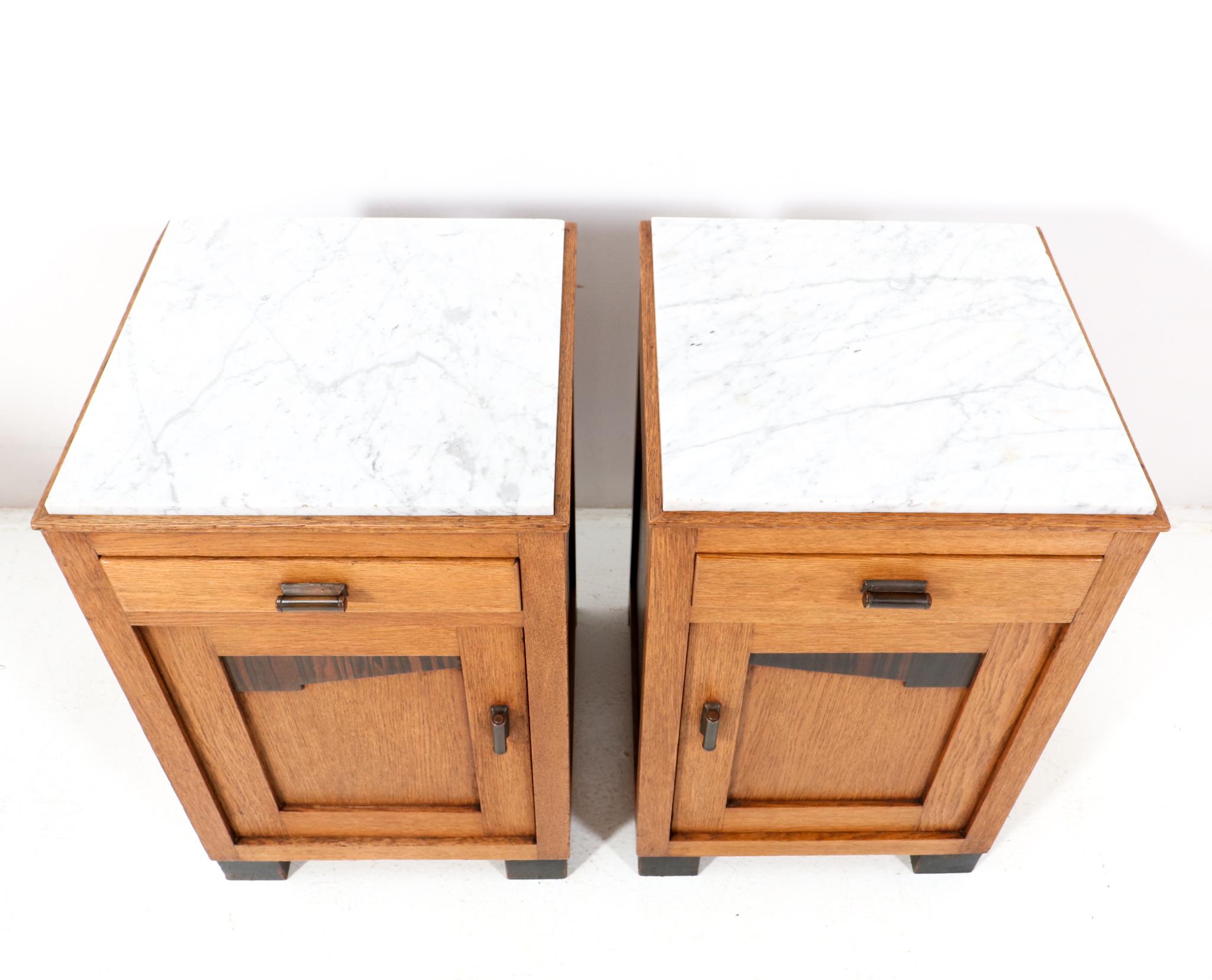 Amazing and rare pair of Art Deco Amsterdamse School nightstands or bedside tables.
Striking Dutch design from the 1920s.
Oak with macassar ebony elements in the doors.
Original stylish brass handles on doors and drawers.
The white marble tops are
