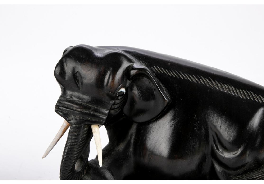 A Pair of Ebonized hardwood Elephants in stylized form. Likely Japanese, the Pair stand with heads turned and striding forward with one front leg extended. Mounted on intricately carved stands. They have glass eyes and plastic or resin removable