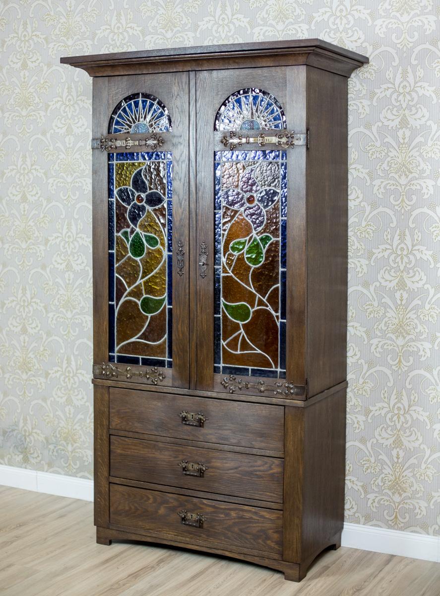 A piece of furniture, circa 1930, made in oaken wood.
The lower section in the form of a dresser with three drawers.
The top is two-piece, crowned with a prominent, profiled molding.
The door panels with stained glass, closed at the top with an