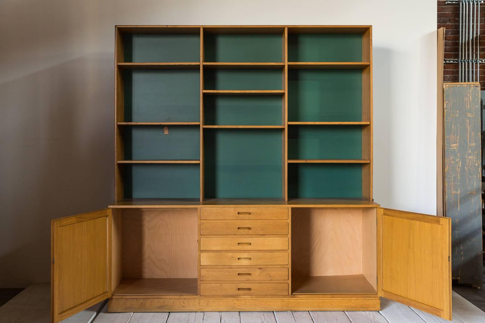 Two-piece midcentury oak cabinet with green back wall. Top piece has adjustable shelves, bottom portion includes a mix of drawers and two doors. Recessed pull details.