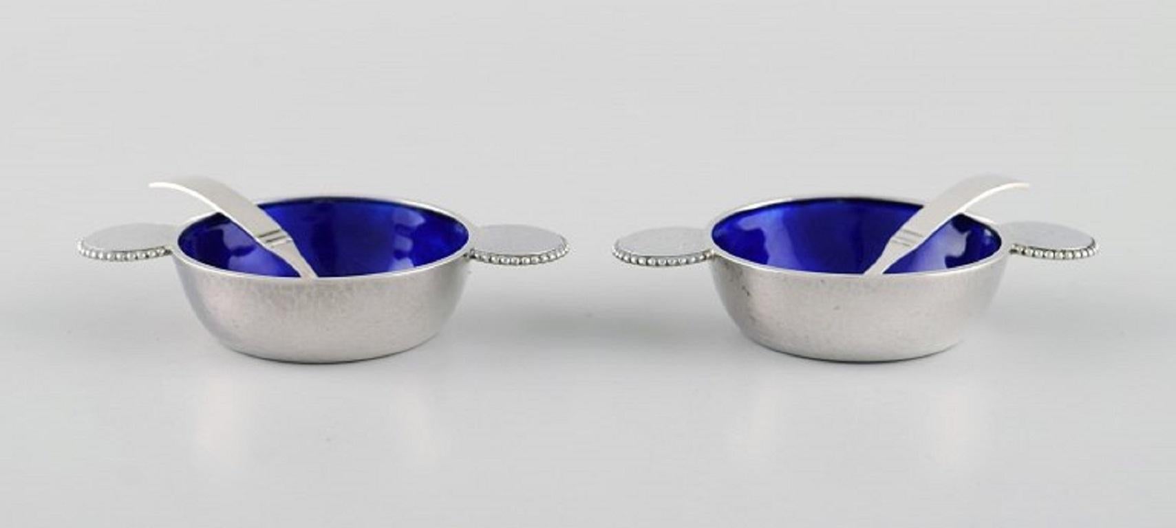 Zwei seltene Salzgefäße von Evald Nielsen aus Sterlingsilber mit königsblauer Emaille und Perlenrand. 
Löffel eines dänischen Silberschmieds. 
1920/30's.
Das Salzgefäß misst: 7.8 x 1,5 cm.
Länge des Löffels: 5,5 cm.
In ausgezeichnetem