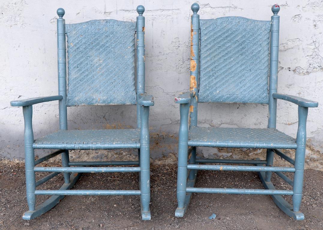 Two rustic Americana style porch rocking chairs with turned wood frames, and woven wicker seats and backs. Bothy are sturdy and solid, both show significant wear to the paint finish, including peeling and loss to paint (blue grey). Offered and