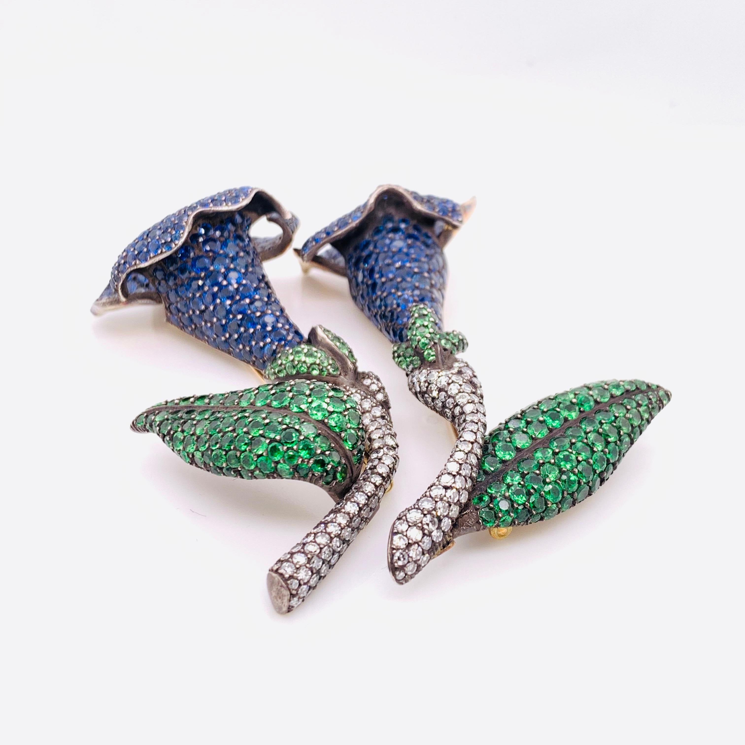 Two sapphire, tsavorite garnet and diamond gentian brooches, mounted in silver-upon-gold, sapphire flower with tsavorite garnet leaves and diamond set stems, with articulated petals and leaves. Measures 70mm and 65mm in length. Estimated total