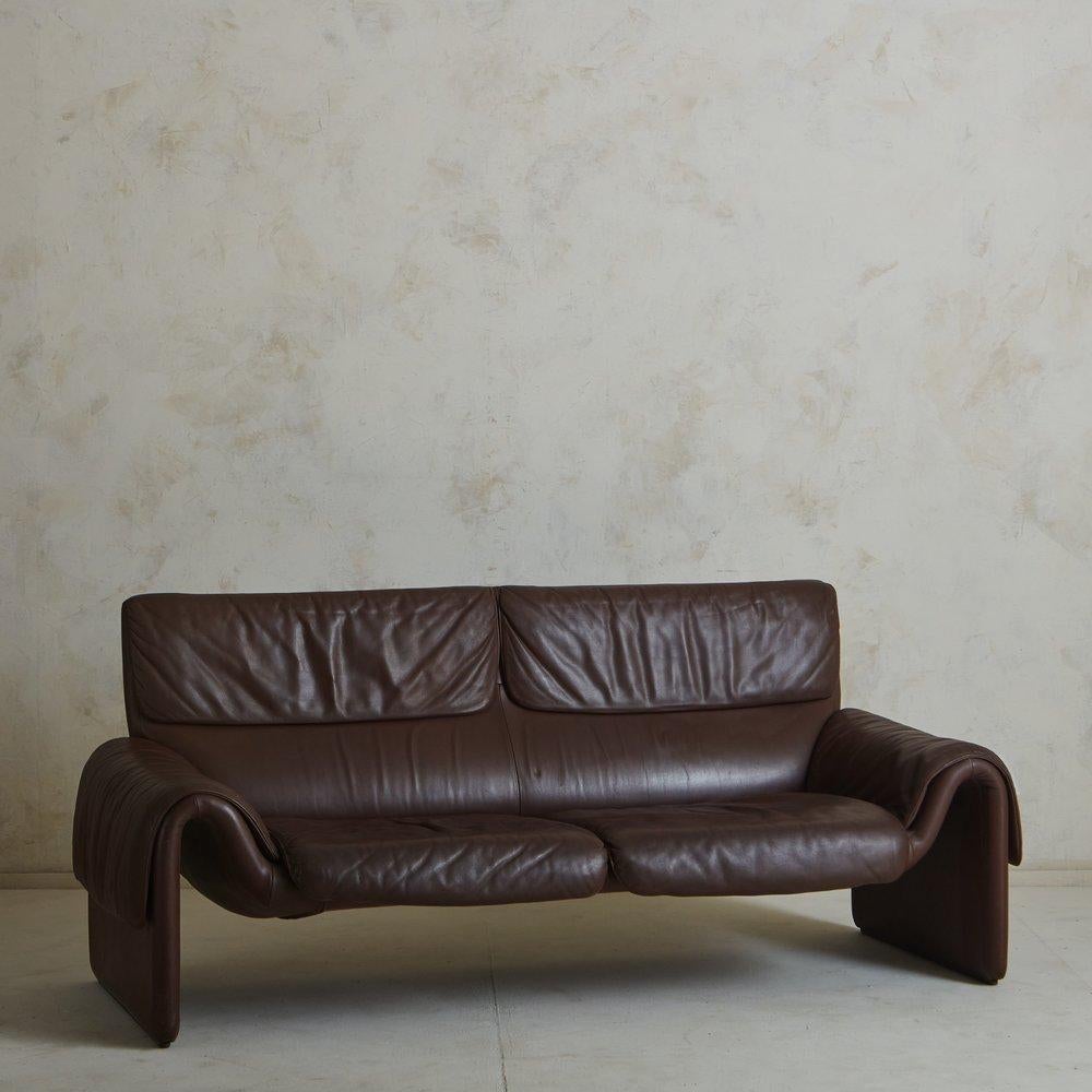 A 1980s Swiss two-seat DS-2011 sofa by De Sede. This sofa has a steel frame and features original patinated leather upholstery with stitch detailing. It has a sculptural silhouette with curved arms and elegant overlaid leather arm rests. Retains ‘De