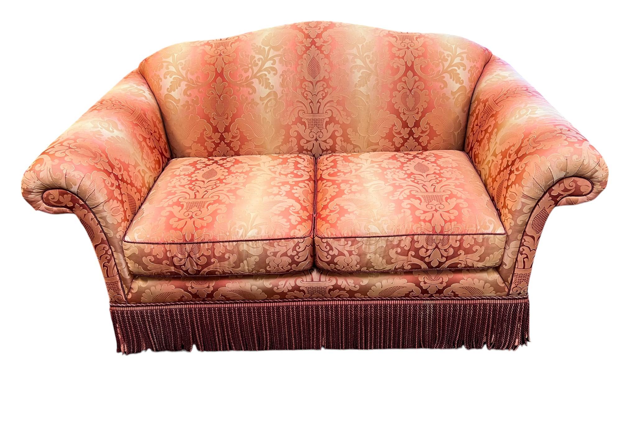 Experience the timeless elegance of this Kravet Furniture two-seat sofa,  upholstered in a satin damask, it features a graceful camelback, classic rolled arms, and contrast welting. A decorative fringe along the base completes the look. The sofa