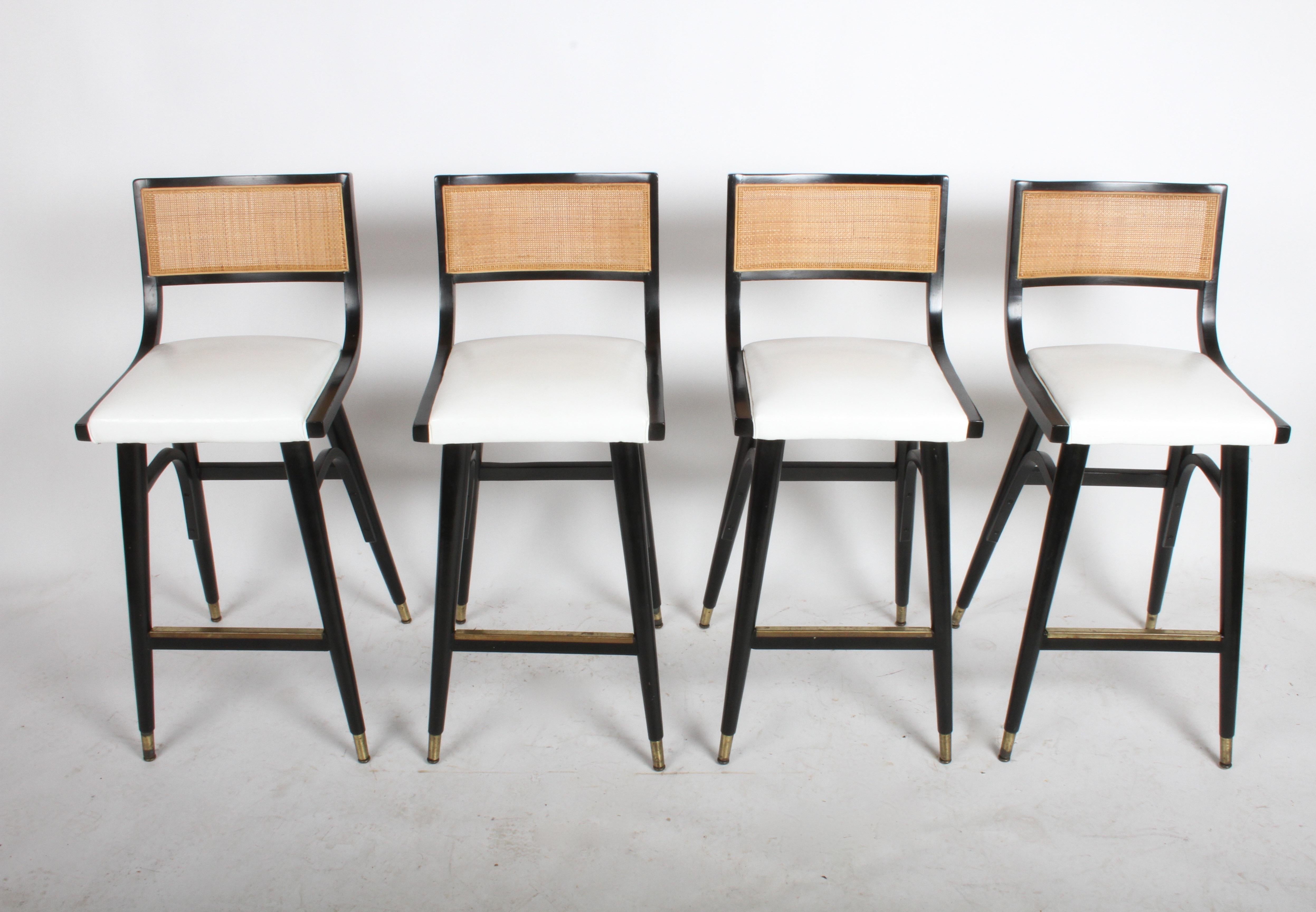I have four matching bar stools Mid-Century Modern bar stools in the style of Edward Wormley for Dunbar with caned backs, black lacquered frames, brass sabots, white vinyl seats and brass covered footrest. All original condition, overall very nice,