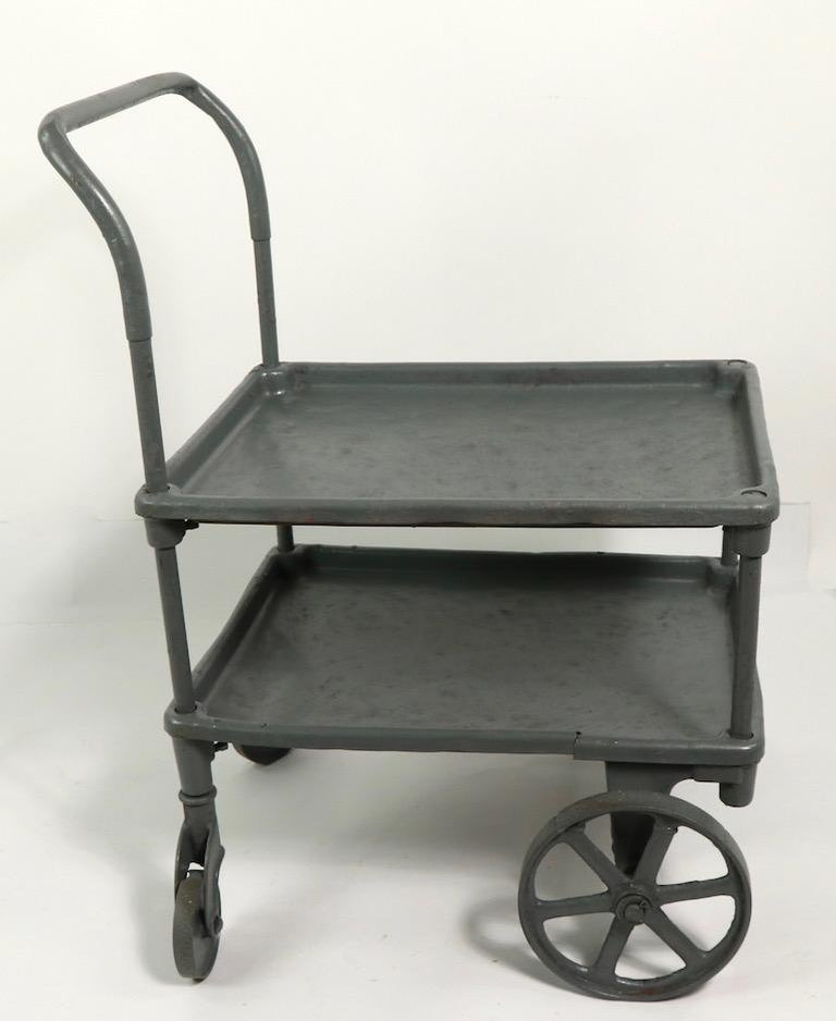 Early industrial design cart having two shelves, and an extended handle, on exceptional cast wheels. This example is currently in later battleship grey paint finish. Clean and ready to use condition.
Slight split in lower shelf, please see