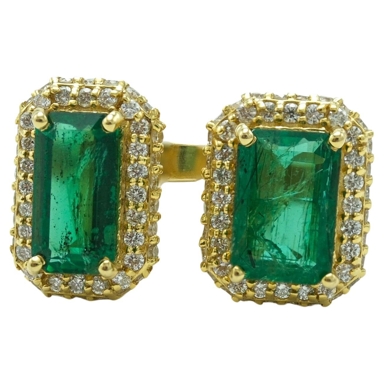 This ring is elegantly constructed from 18-karat yellow gold and showcases two emerald-cut emeralds with a total weight of 3.5 carats. The gemstones radiate a deep, vibrant green, and are framed by a halo of round diamonds totaling 0.5 carats. The