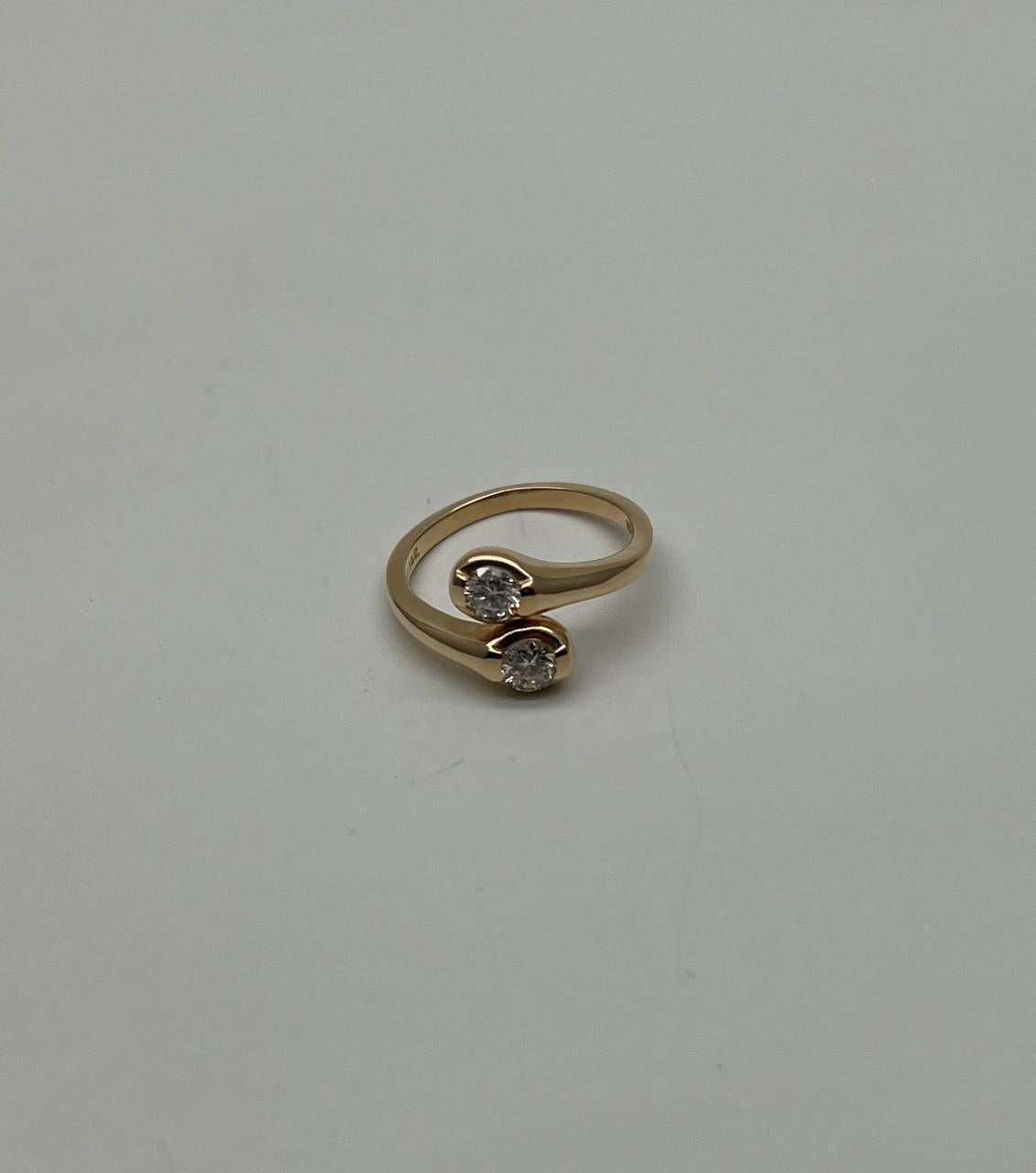 Cette bague exquise présente un anneau d'or élégant qui s'enroule gracieusement autour du doigt. Le design met en valeur deux diamants ronds étincelants, chacun serti dans une lunette lisse et polie. L'anneau ouvert ajoute une touche de modernité et