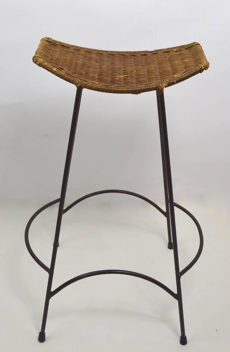 Woven wicker saddle seats on iron rod base, in the style of Arthur Umanoff. Wicker seats show minor loss, normal and consistent with age. These stools will stack for easy storage. Seats 16 L x 10.5 W. Offered and priced individually.
