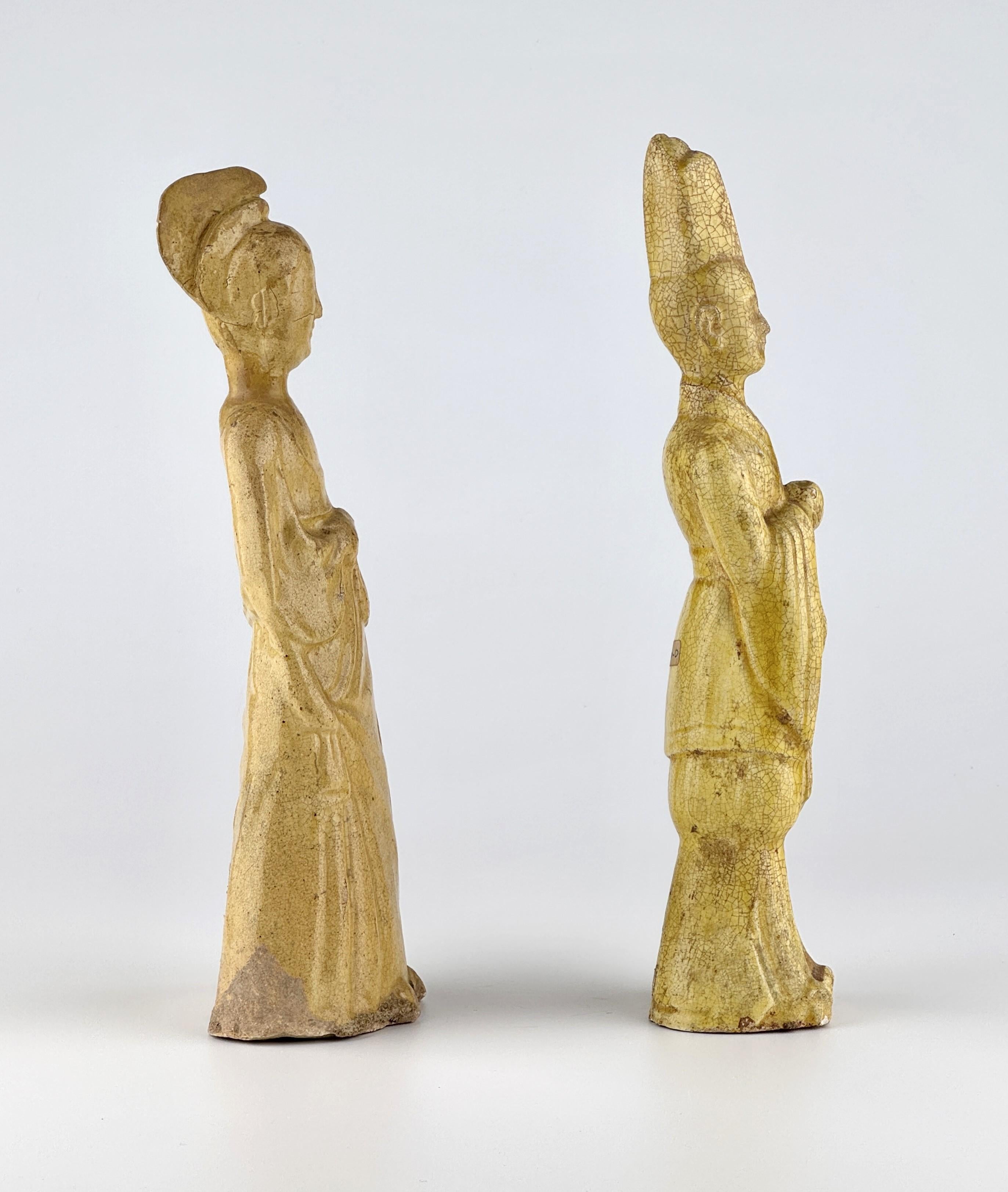 This pair of straw-glazed pottery figures represents a male and female court attendant, both elegantly attired in period fashion. The female figure wears a flowing robe with draped folds and a high-stylized coiffure, while the male figure dons a