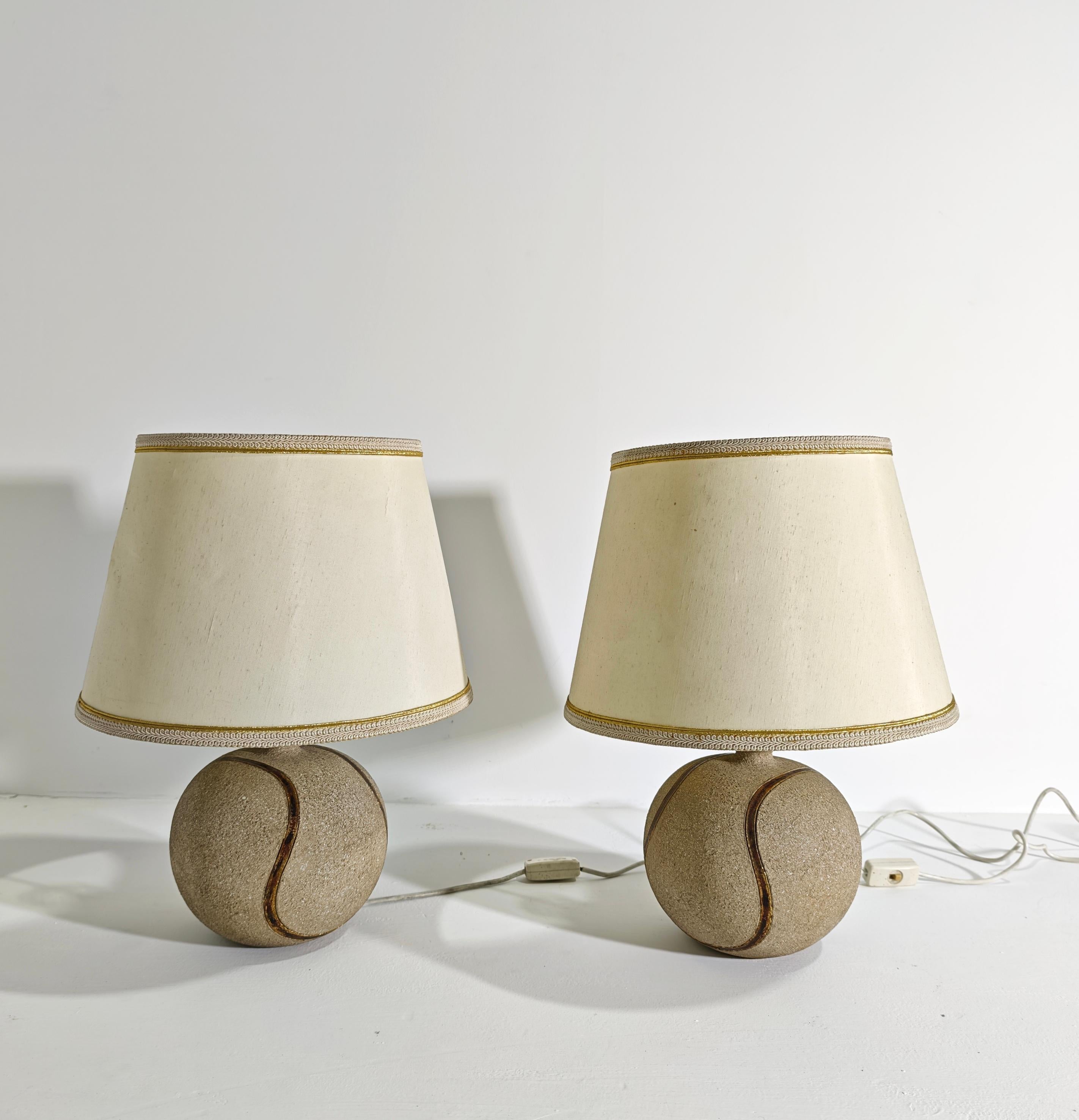 Zaccagnini Ugo. Made in Italy, ca. 1970
Pair of ceramic table lamps with a ground stone effect. Circular body with a narrow neck and a flat, everted spout; decorated with a shaped gold ribbon. Ivory-colored fabric lampshades. Manufacturer's mark on