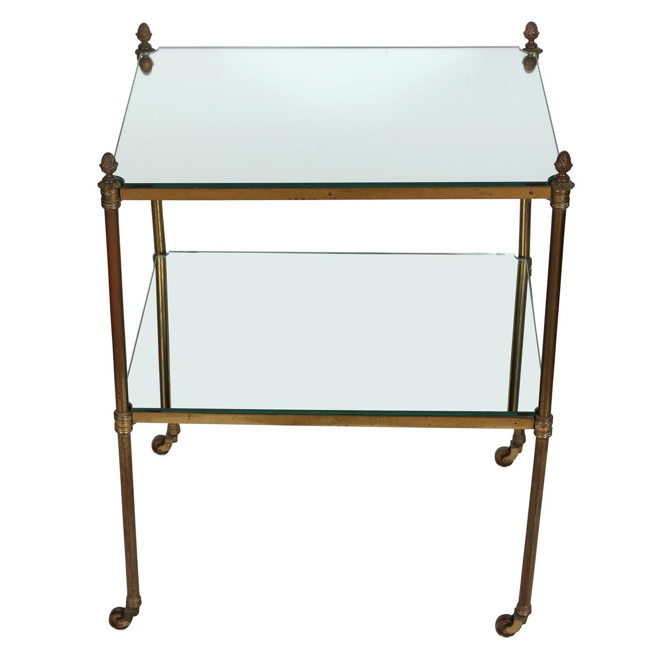 A vintage brass two tier side table with mirrored shelves.  Finished with pinapple finials at each corner and sitting on casters, this elegant Hollywood Regency style table will be at home in many style interiors. Mirrored shelves are new.  Natural
