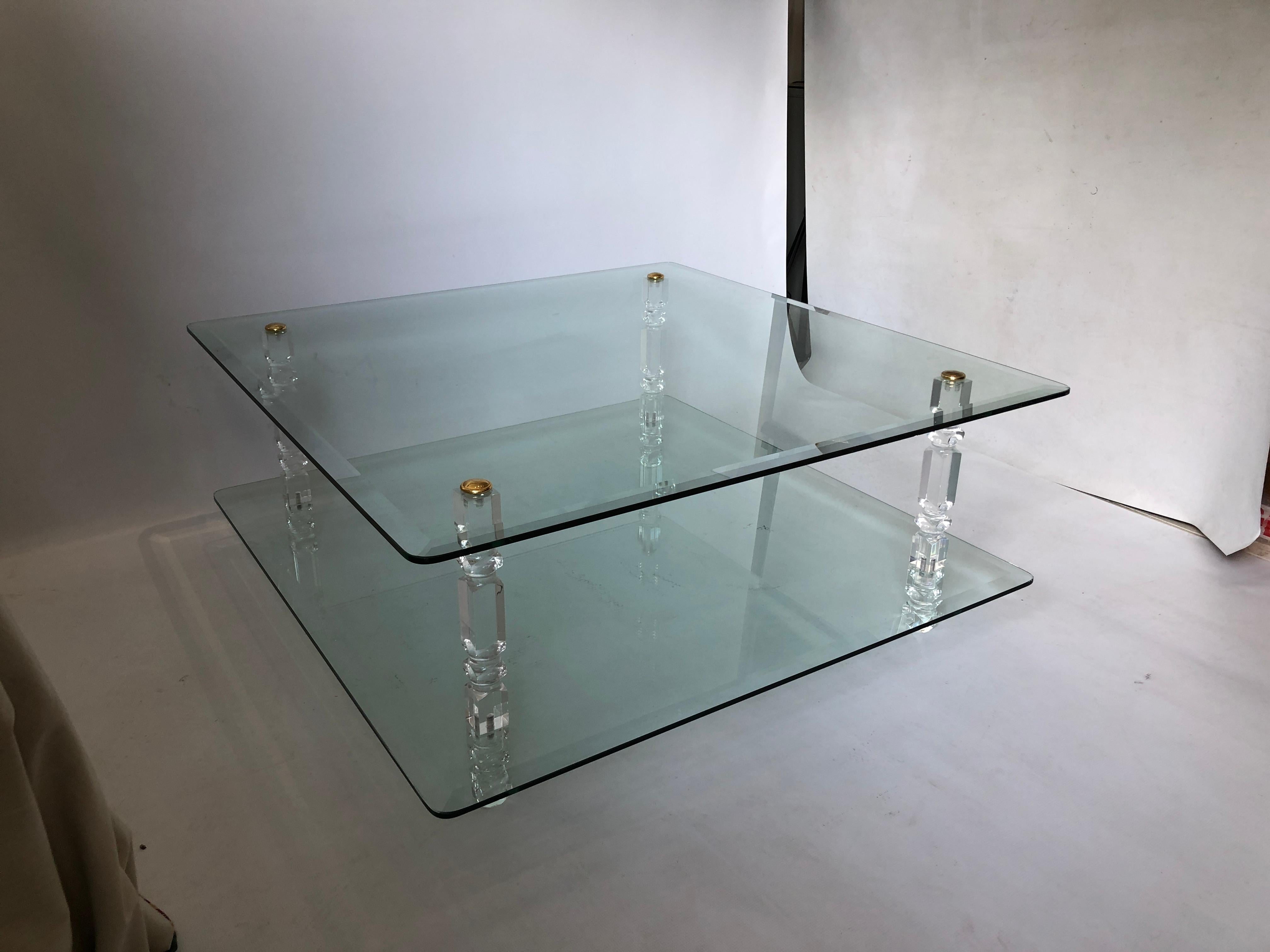 This large, two-tiered coffee table is composed of two square sheets of bevelled, reinforced glass, gently curved at the corners. These are held in place by four ornate lucite columns, with a brass disc screw at the top to complete the piece. The