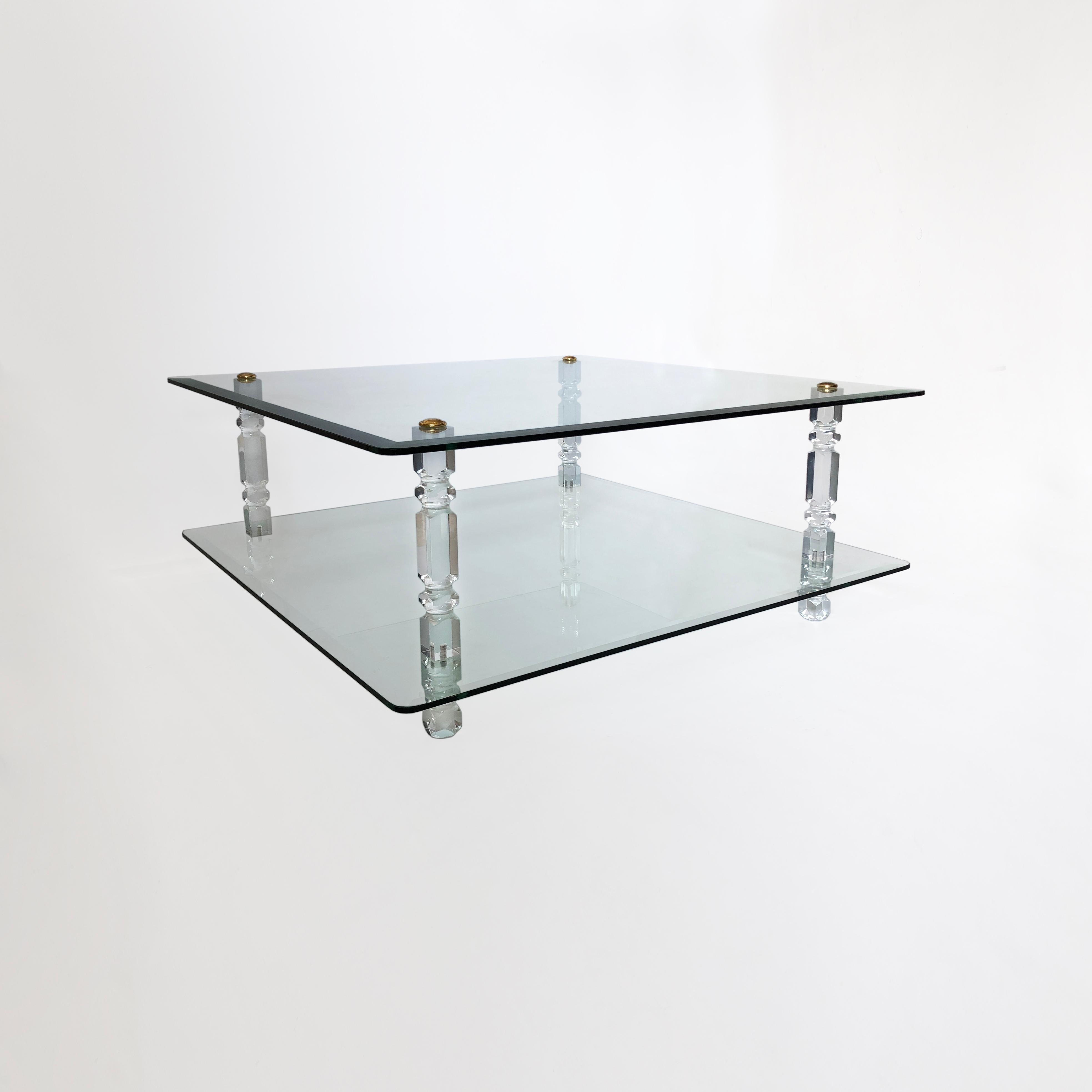 This large, two-tiered coffee table is composed of two square sheets of bevelled, reinforced glass, gently curved at the corners. These are held in place by four ornate lucite columns, with a brass disc screw at the top to complete the piece. The