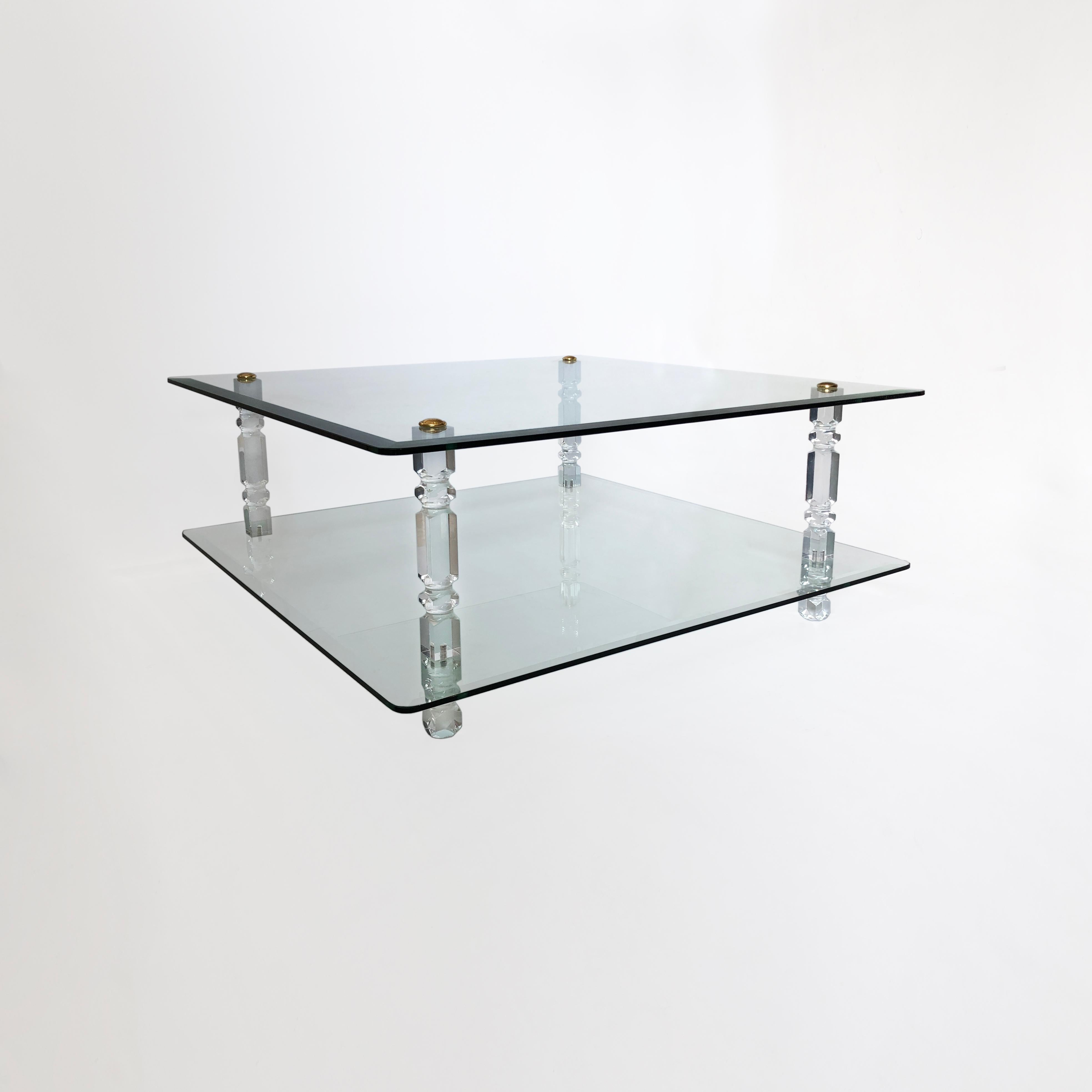 Cette grande table basse à deux niveaux est composée de deux plaques carrées de verre armé biseauté, légèrement incurvées aux angles. Celles-ci sont maintenues en place par quatre colonnes en lucite ornées, avec une vis à disque en laiton au sommet