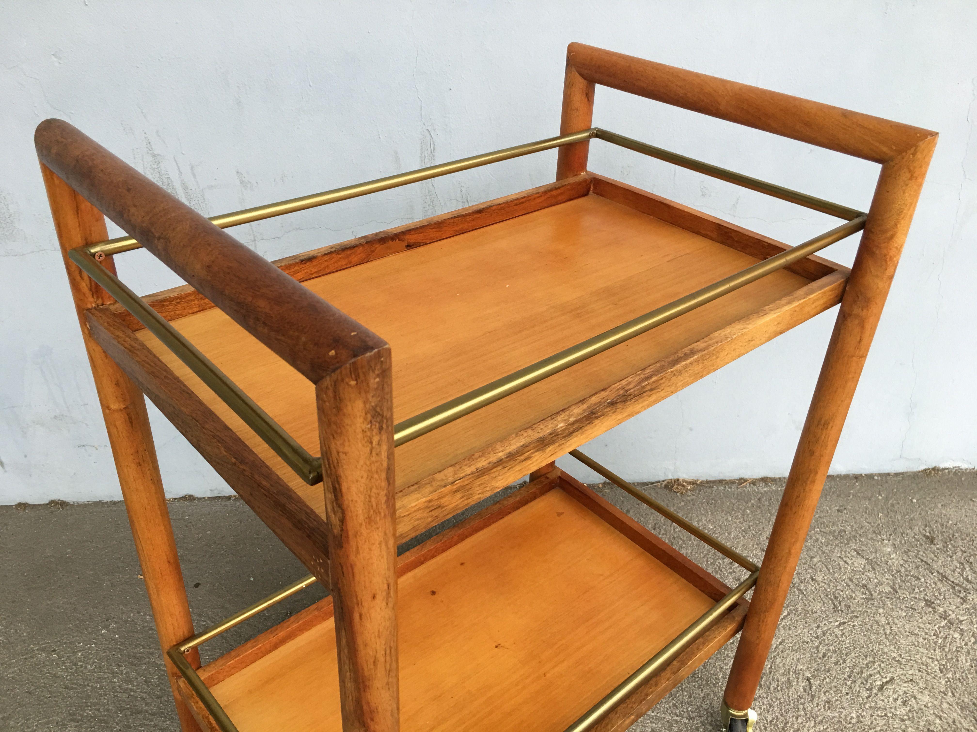 Dark blond two-tier midcentury bar cart with round-shaped side frames. The cart features brass railings with four brass coaster wheels, two lock to keep cart from moving. Reminiscent of the design styling made popular by the Heywood Wakefield