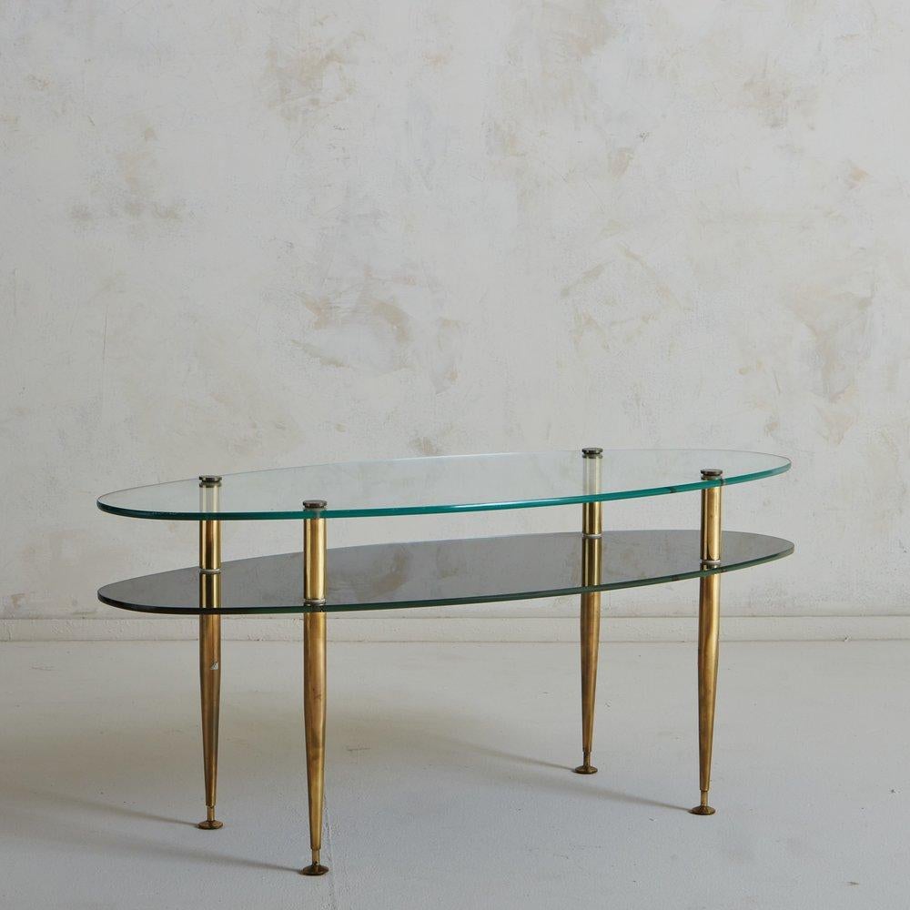An elegant French coffee table featuring an oval glass tabletop with a mirrored glass lower shelf. This piece has tapered patinated brass legs with petite ball feet. The legs screw into the tabletops, allowing for complete disassembly. Sourced in