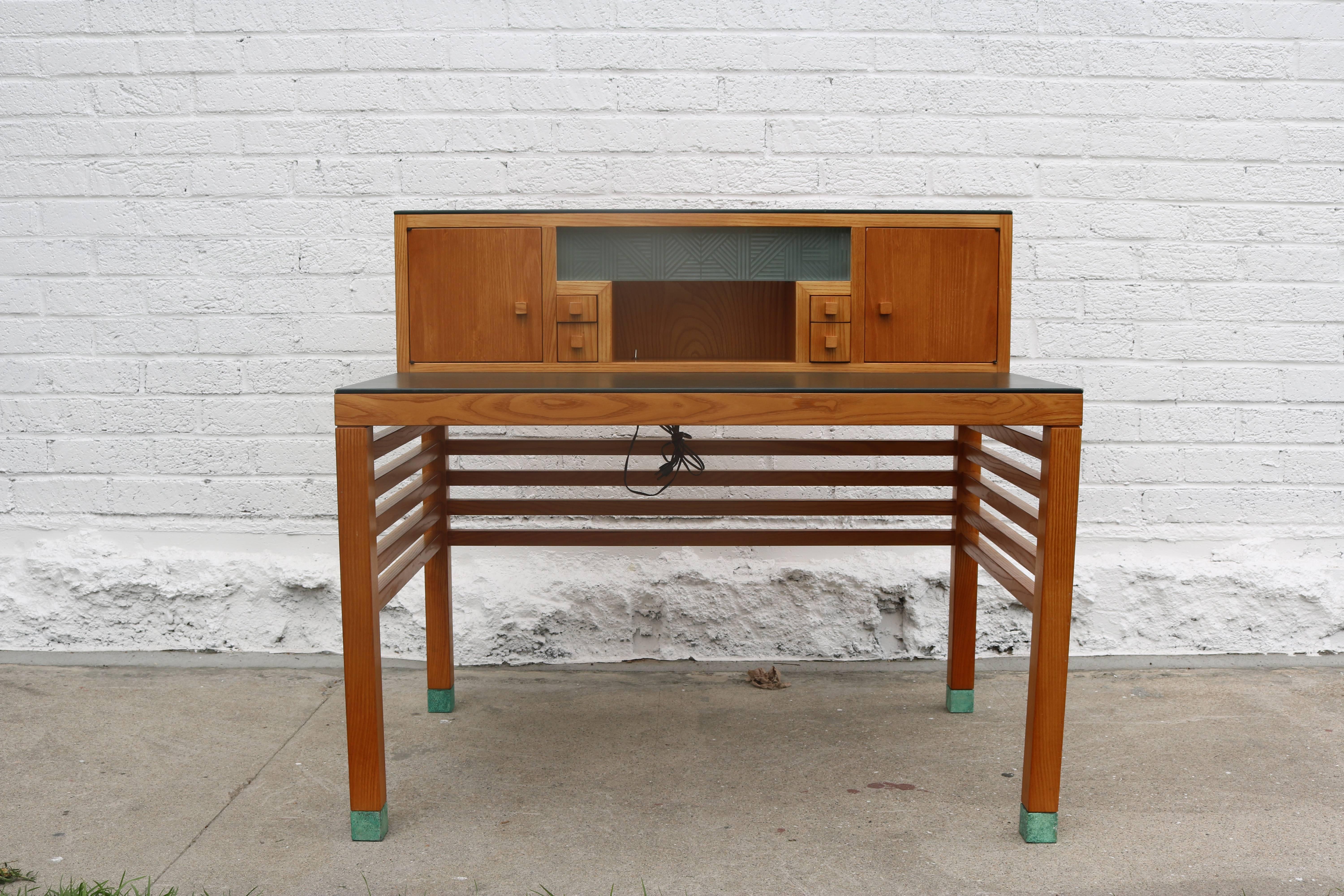 This desk by Steven Holl for Pace has two levels with dark green leather on the surface of each. There is an acid etched glass panel above a small cubby - on either side are two small drawers perfect for pens as well as small doors on either side.